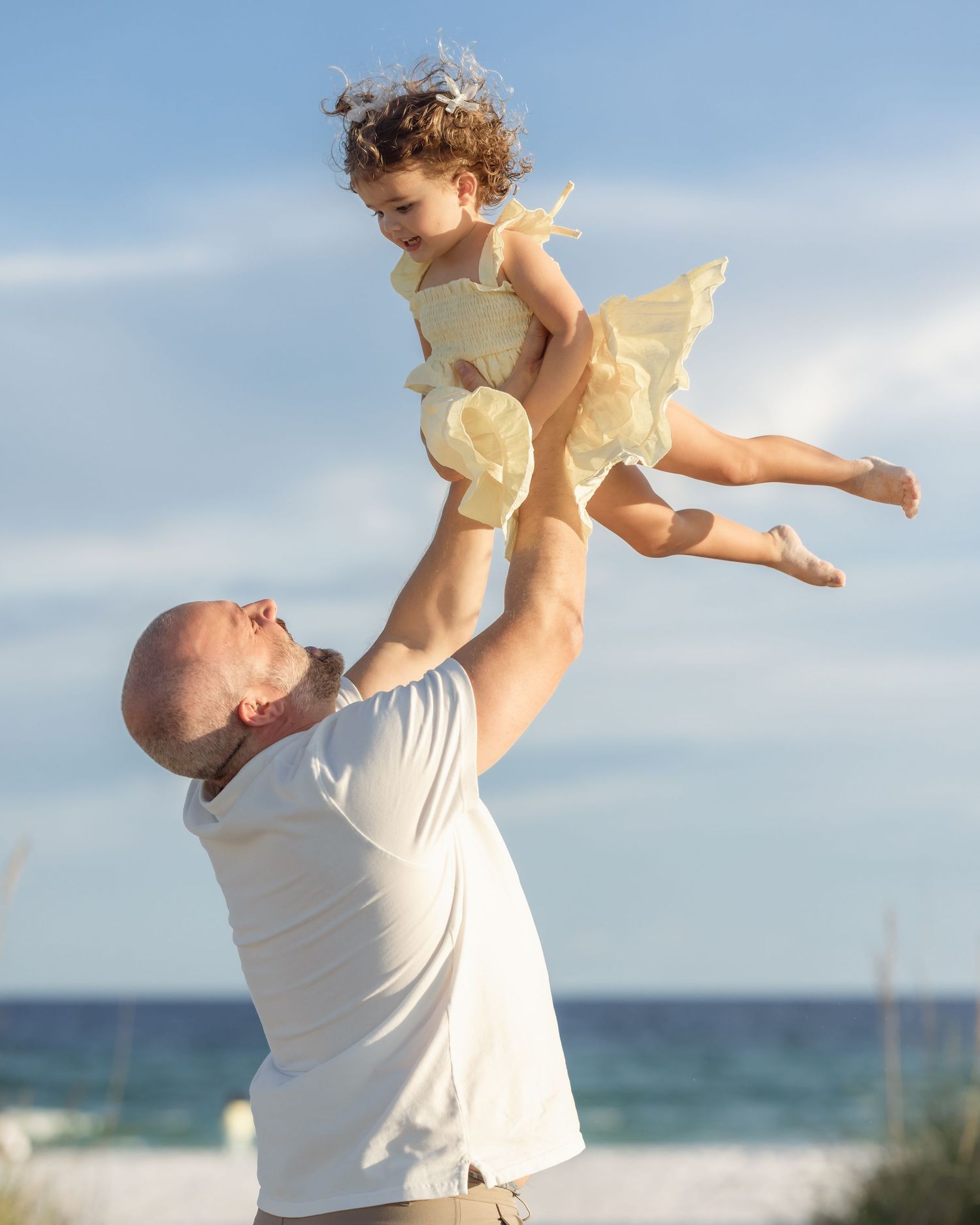 Man tossing young girl in yellow dress into the air on a sunny beach.