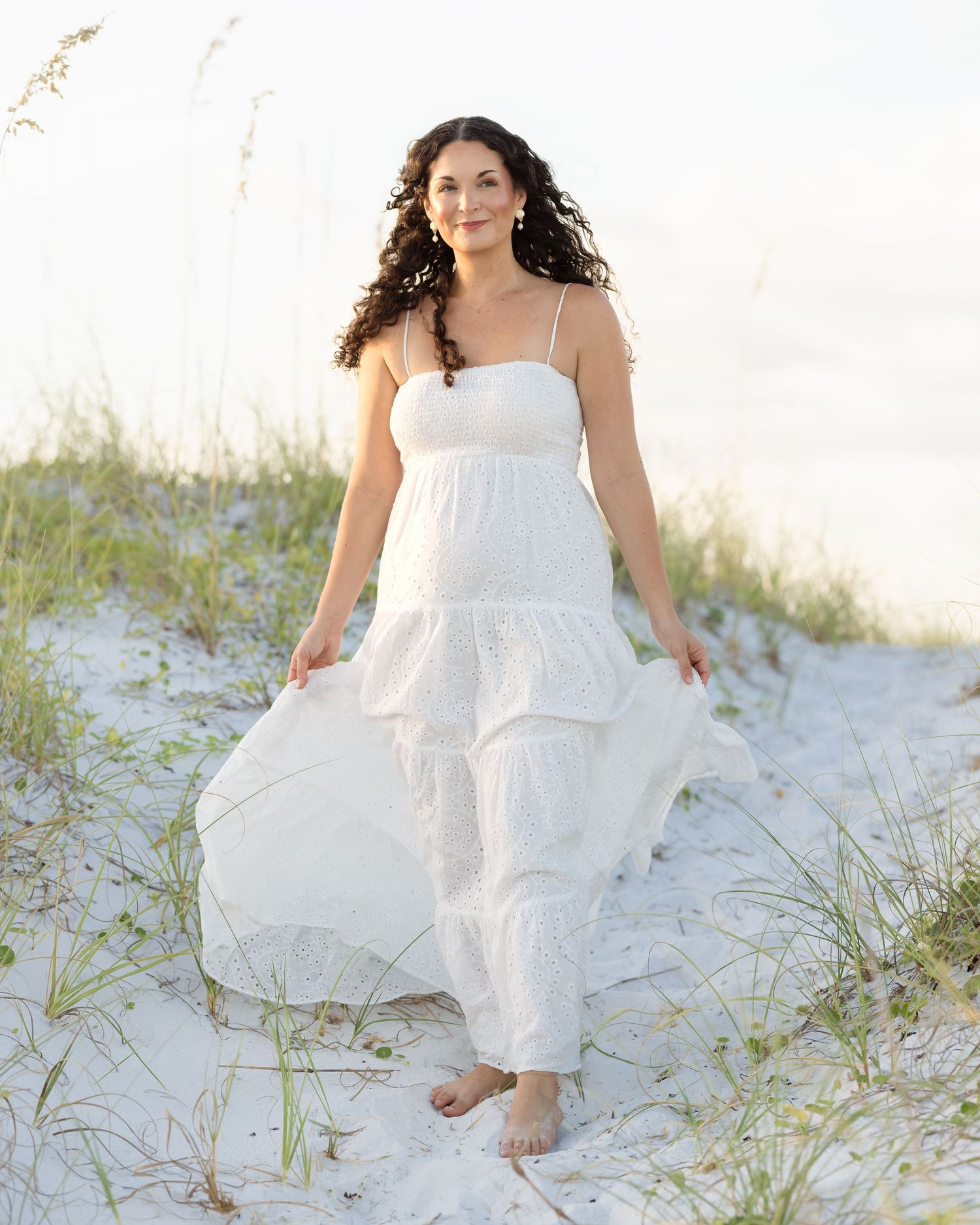 Smiling woman in flowing white maxi dress walking barefoot on white sand beach dunes.