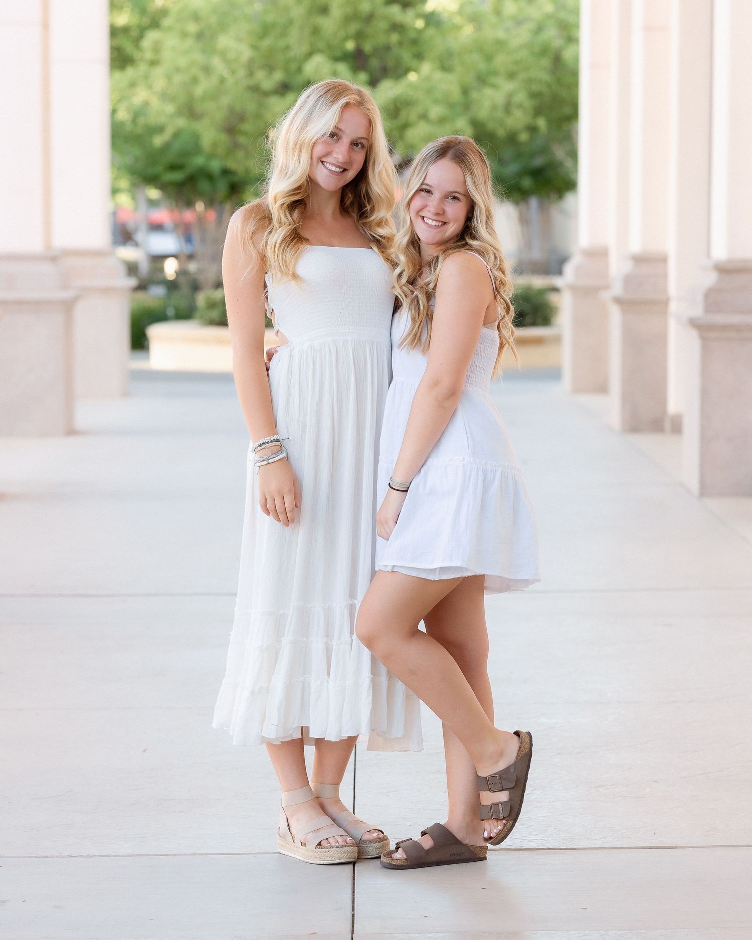 Two young women in white dresses and Birkenstock sandals smiling outdoors near stone columns.