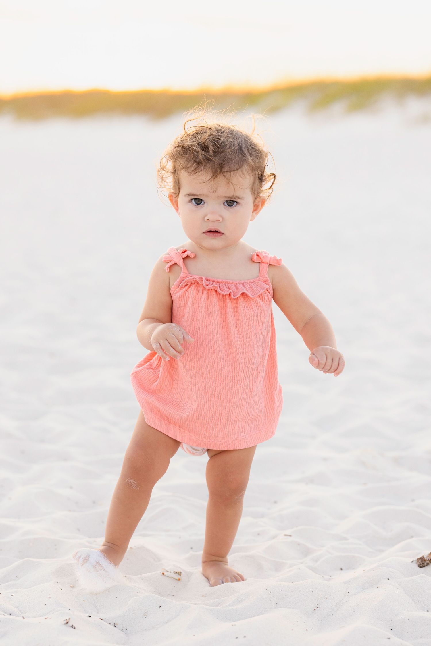Toddler in peach-colored outfit explores soft sand during sunset at the beach.