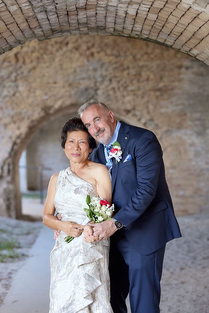A couple in formal wedding attire share an intimate moment against a rustic stone wall backdrop.