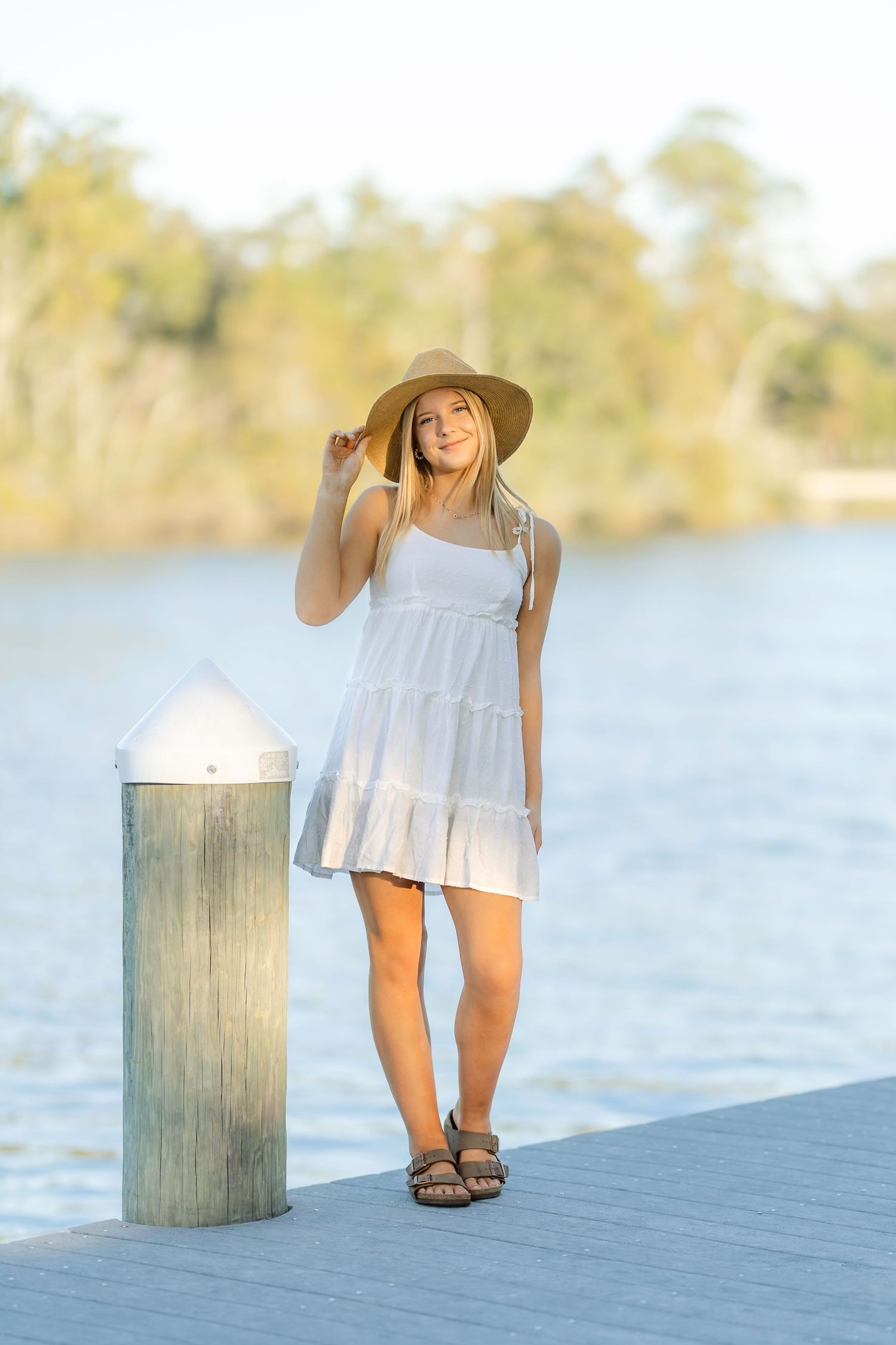 A person in a white flowy dress stands on a wooden dock by a serene lake on a sunny day.
