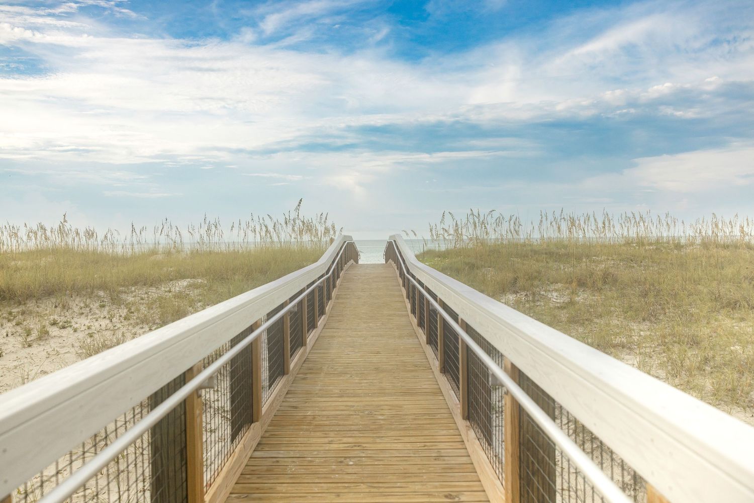 Wooden boardwalk through sandy beach dunes leads to ocean under bright blue sky with white clouds.
