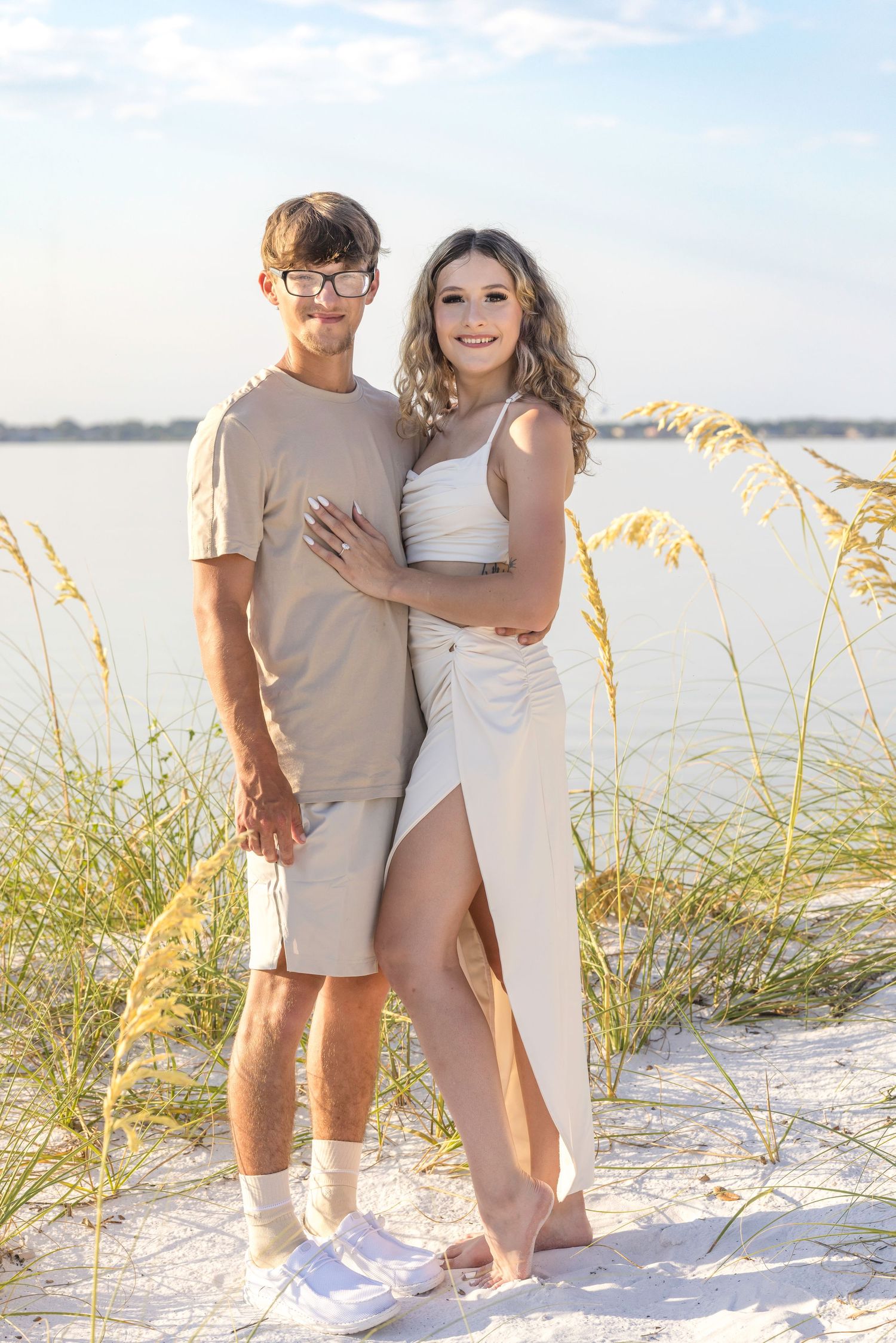 A couple embraces while standing on a white sand beach with golden sea oats and water in the background.