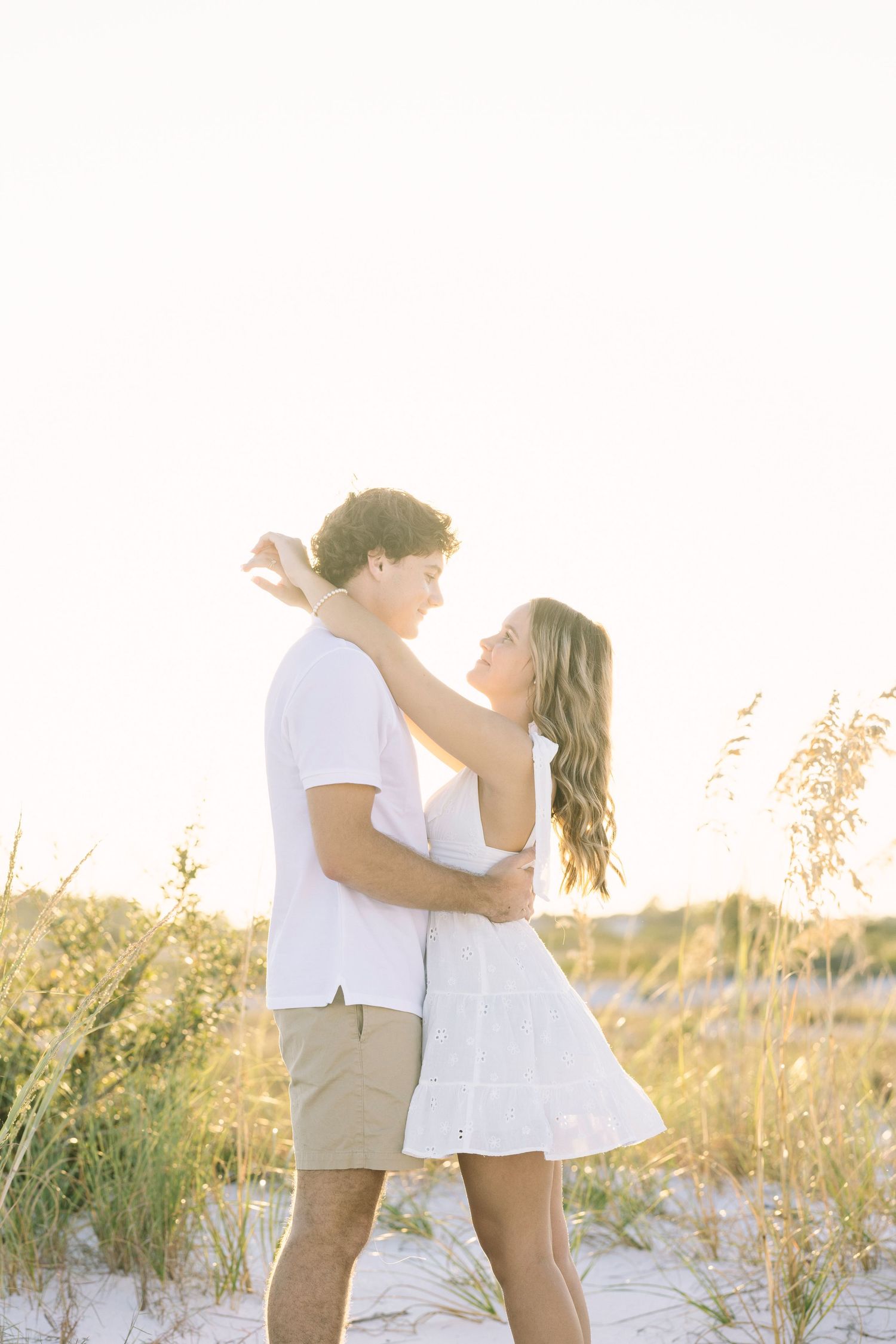 Couple embraces on sunny beach with tall grass during golden hour sunset in white and khaki summer outfits.