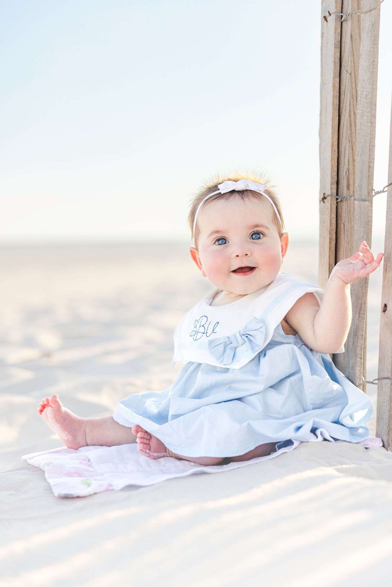 Baby in light blue dress sitting on sandy beach against wooden post in soft natural lighting.