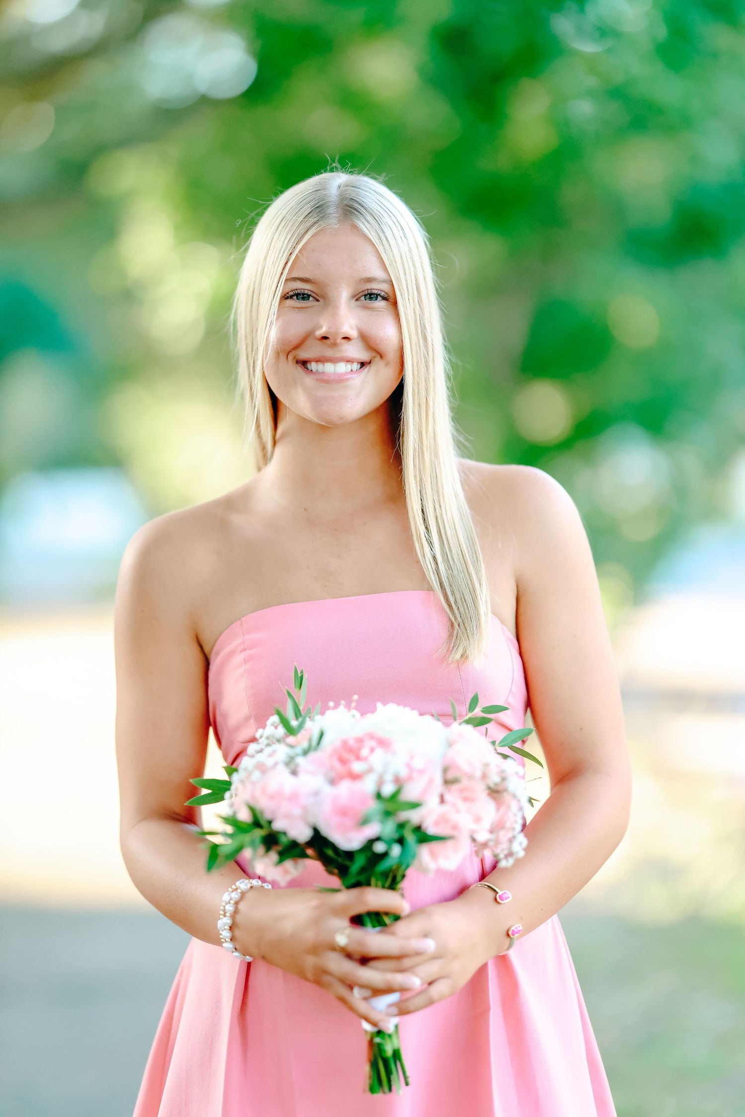 A bridesmaid in a pink strapless dress holds a pink and white floral bouquet while smiling outdoors.