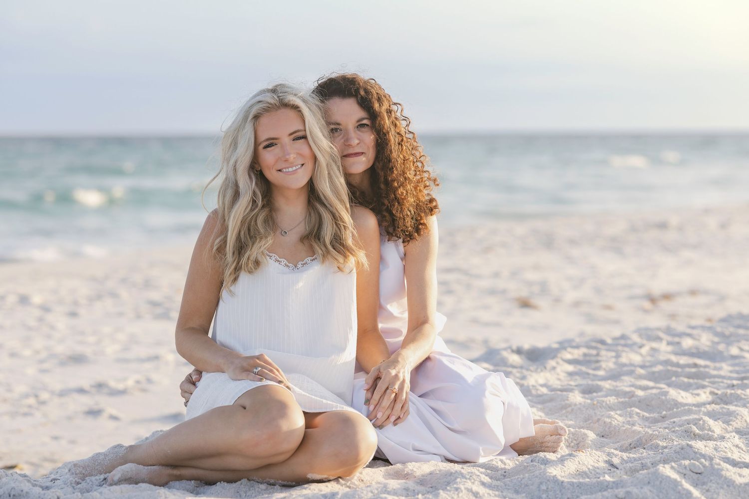 Two women sitting together on a sandy beach, smiling, wearing light pastel outfits with ocean waves in the background.