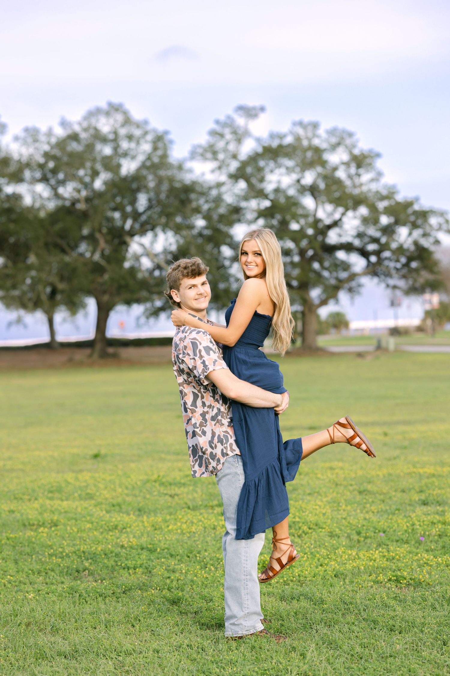 Two people embrace while standing in a grassy park with trees in the background on a sunny day.