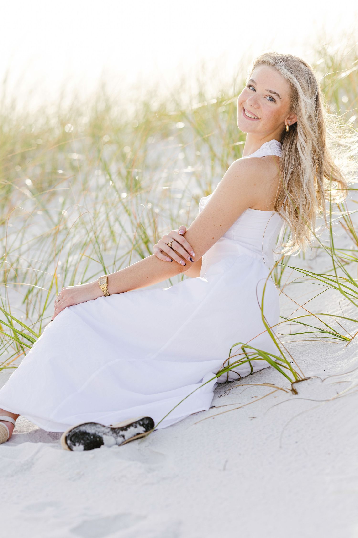 Person in flowing white dress reclines gracefully on sandy beach near sea grass during a photo shoot.