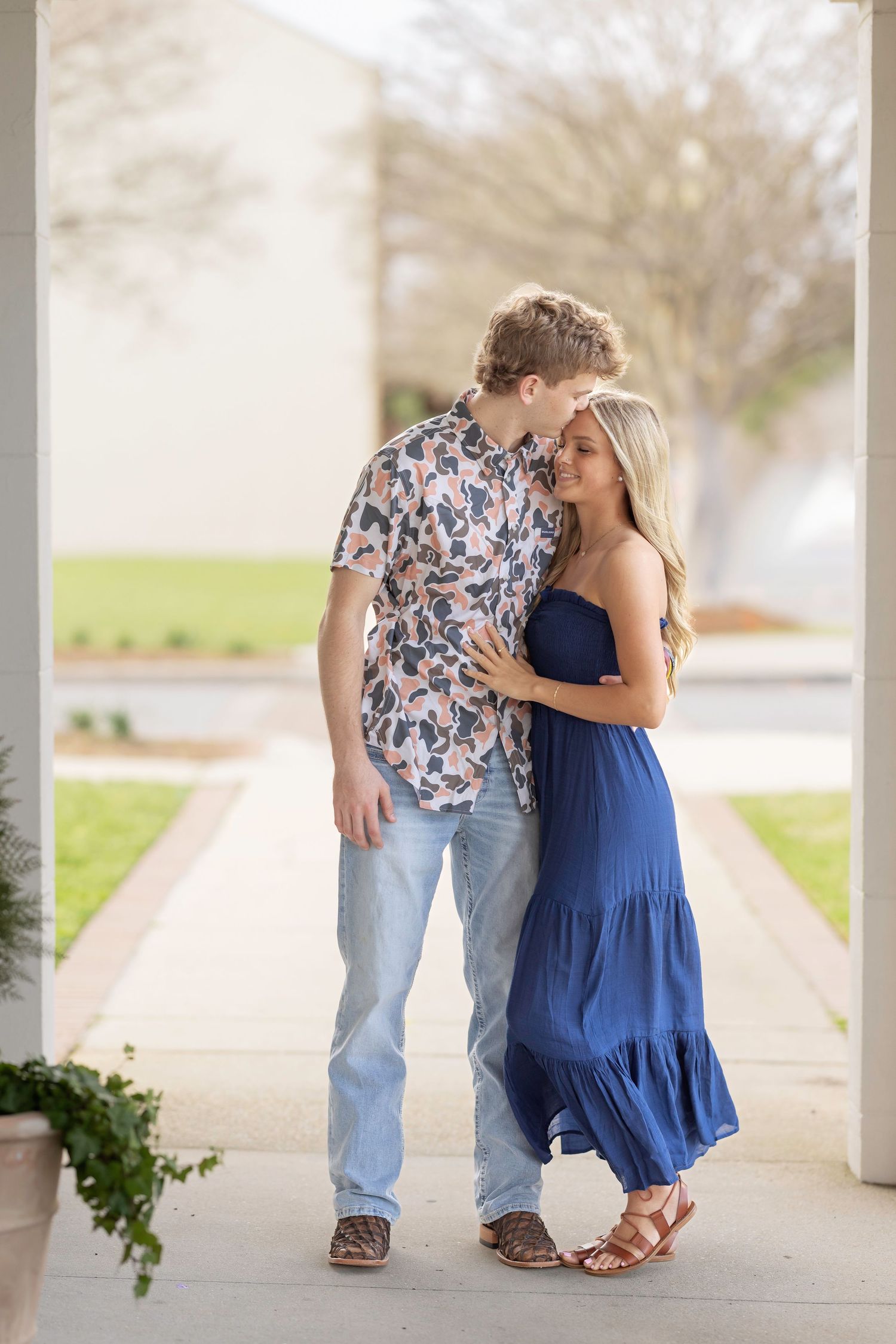 A couple embraces on a covered walkway, with her wearing a navy blue maxi dress and him in a floral shirt and jeans.
