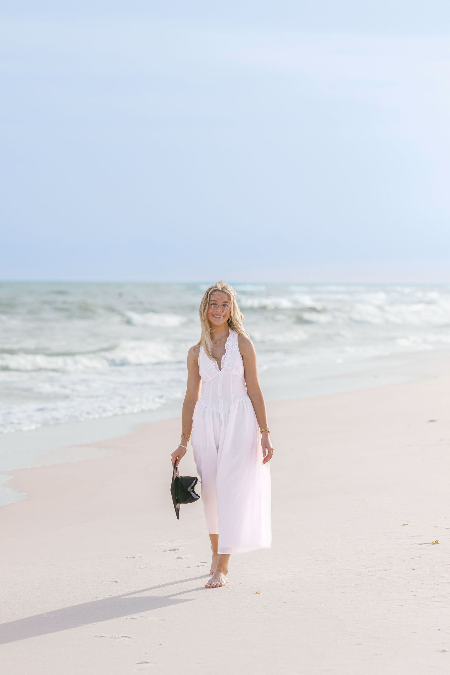 Blonde woman in white maxi dress walking barefoot on sandy beach holding black clutch purse.
