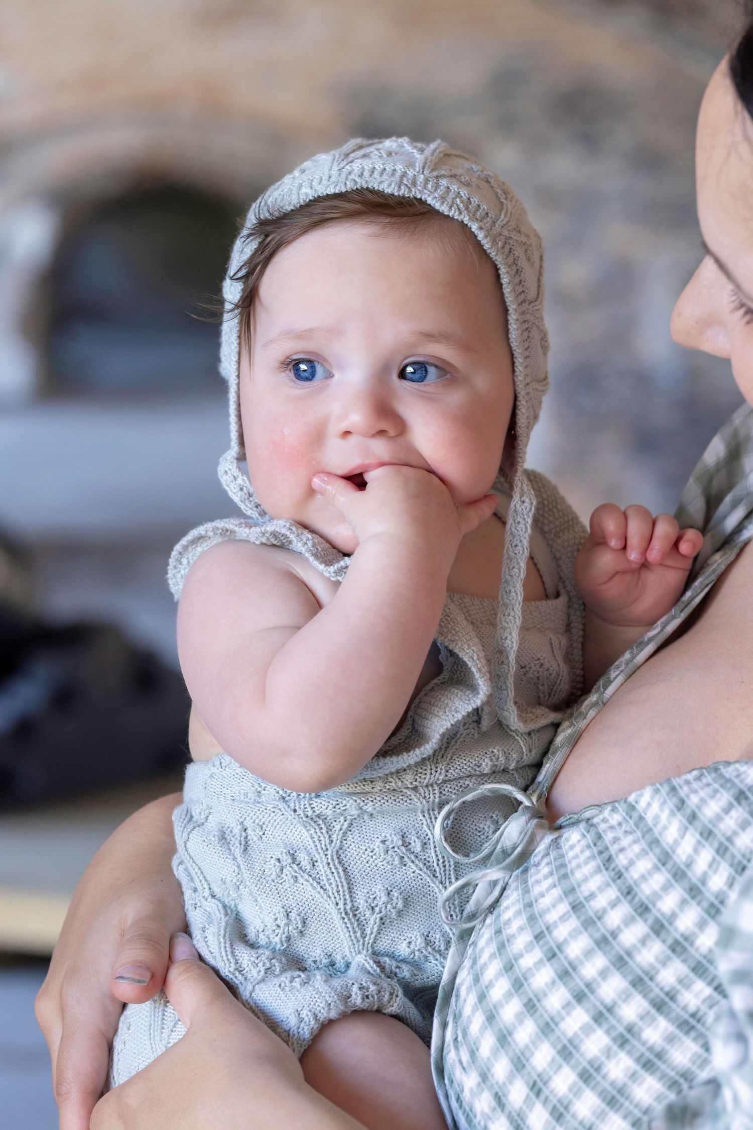 A blue-eyed baby in a gray knit bonnet and romper chews fingers while being held by an adult.