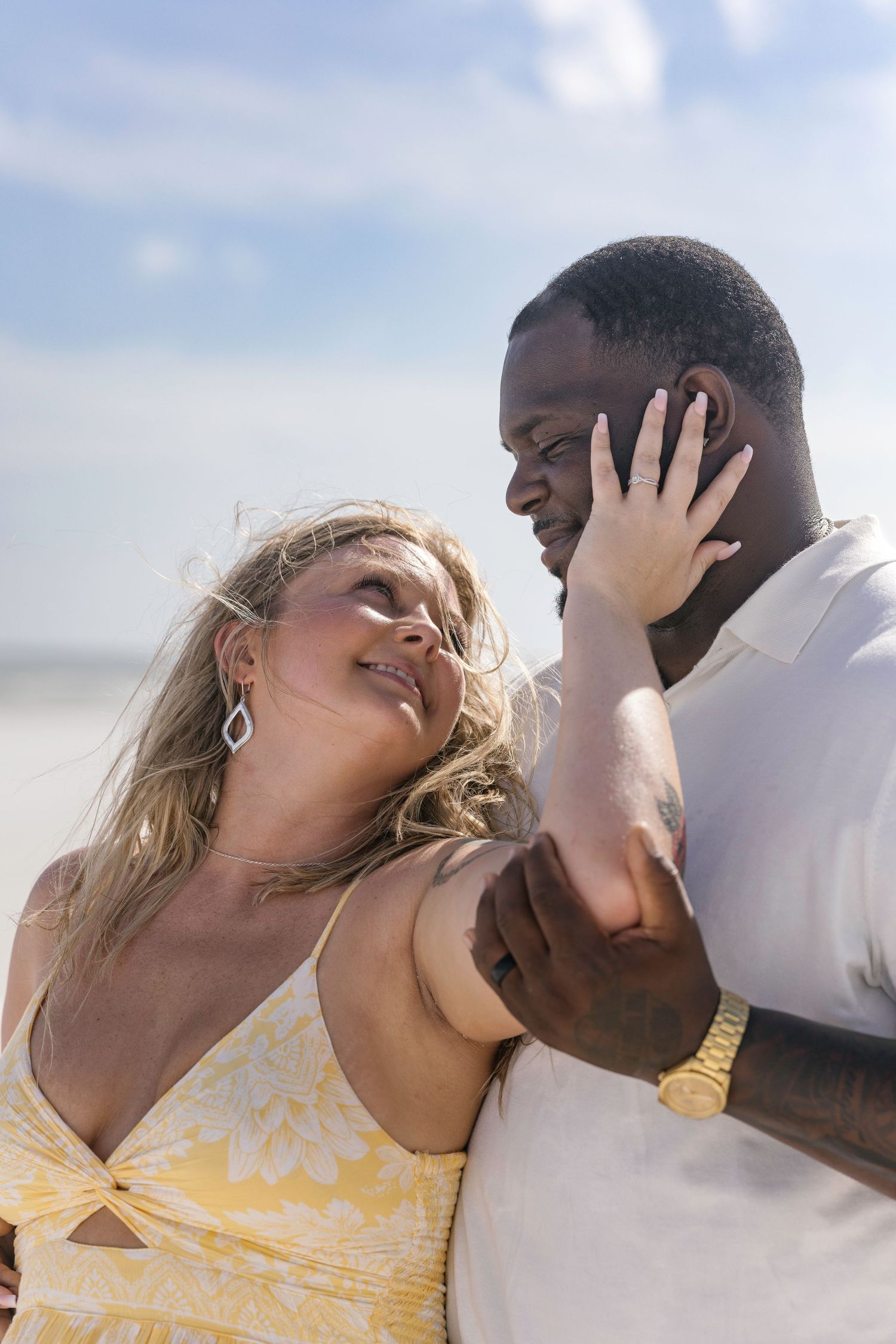 Interracial couple sharing a tender moment on a breezy beach under a partly cloudy sky.