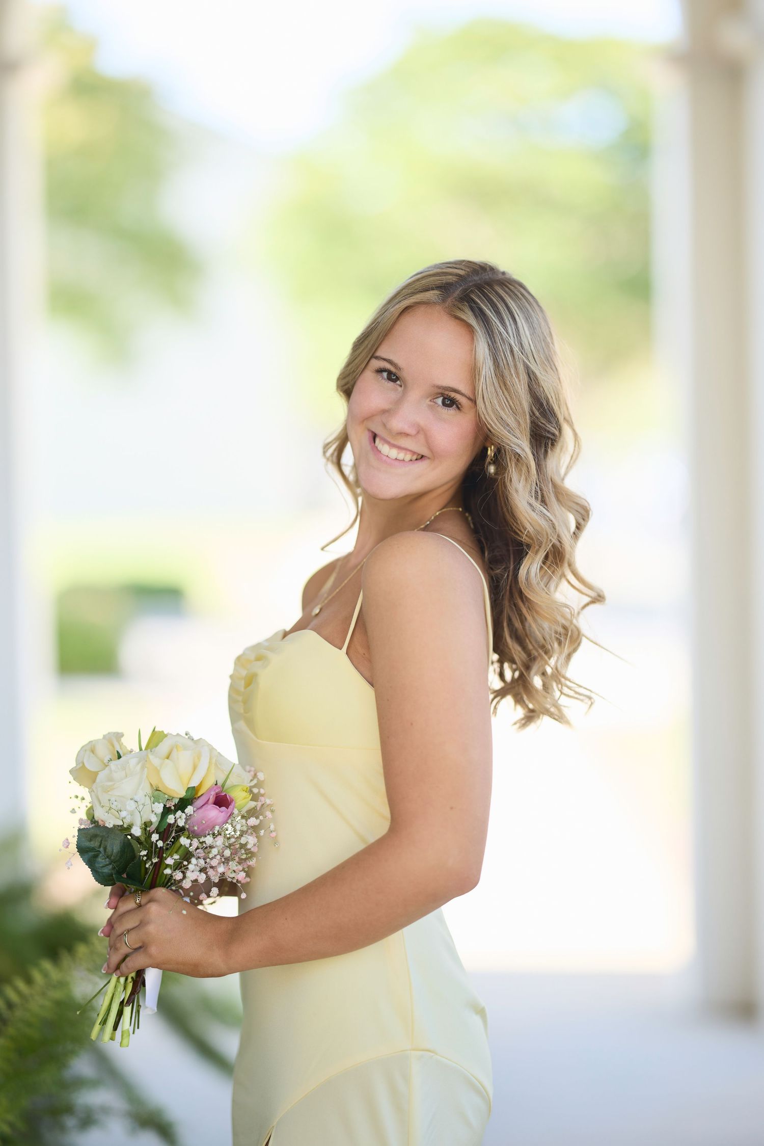 Smiling young woman in yellow dress holding white and pink flower bouquet, posing outdoors near white columns.