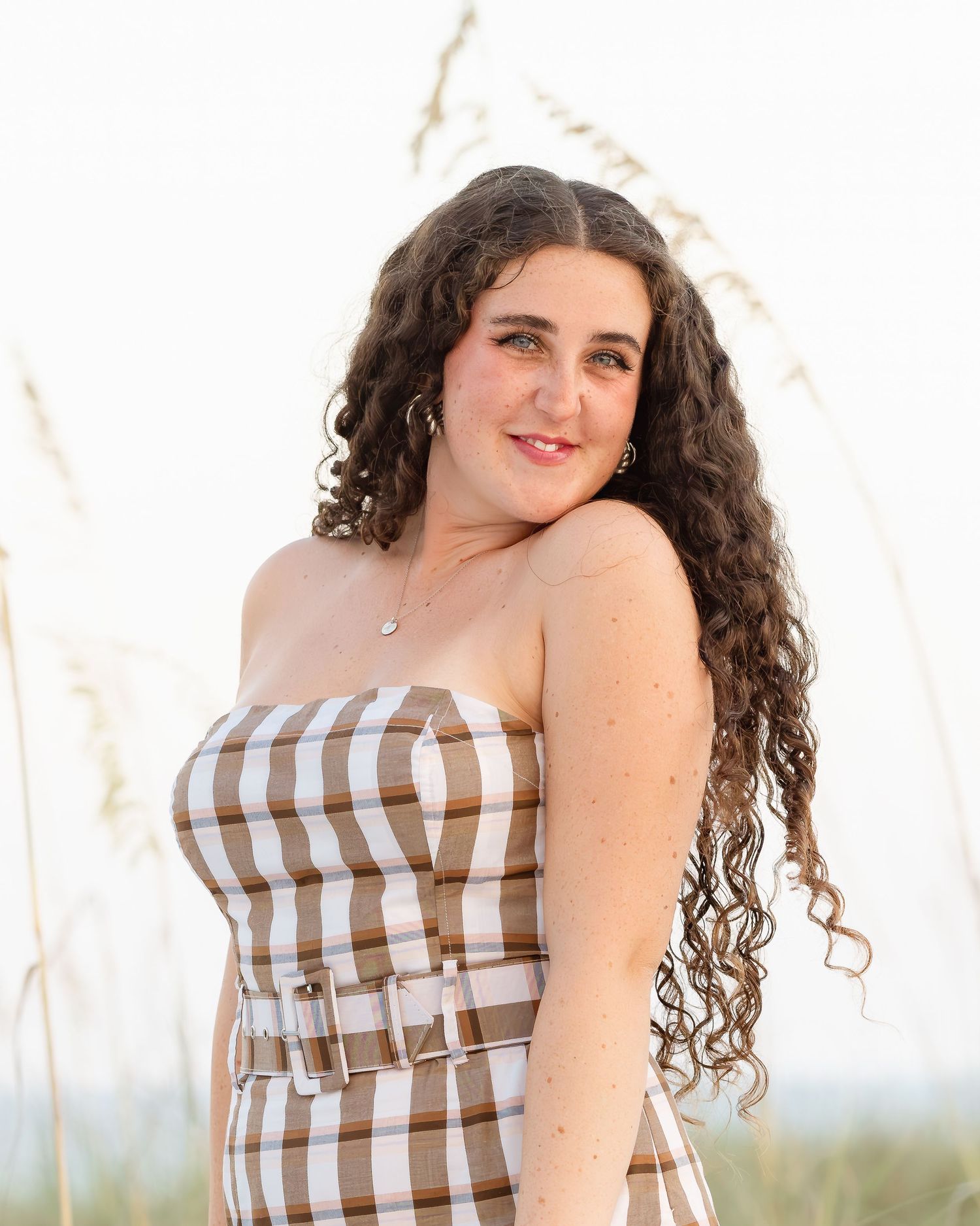 Young woman with curly hair wearing a striped strapless dress poses outdoors near tall beach grass.