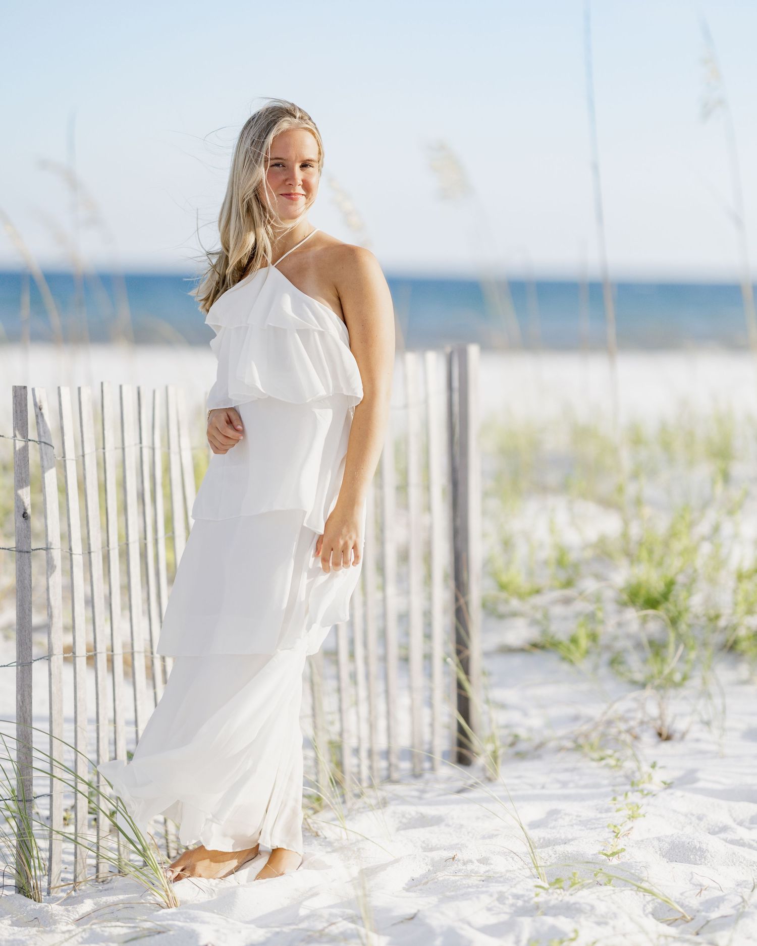 Woman in white maxi dress posing on sandy beach dunes with sea oats and ocean in background.