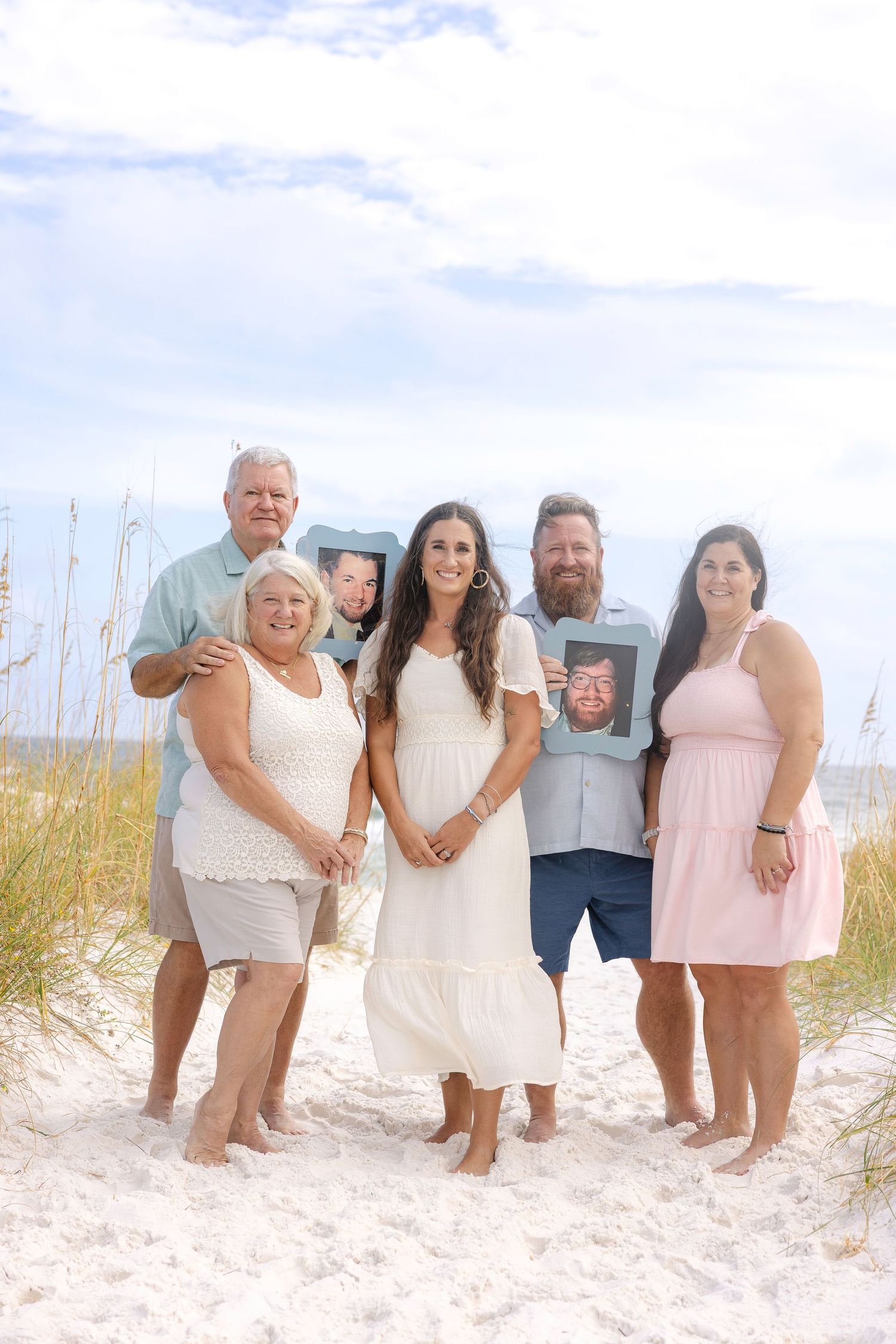 Large family group smiling together on sandy beach with beach grass and blue sky in background.