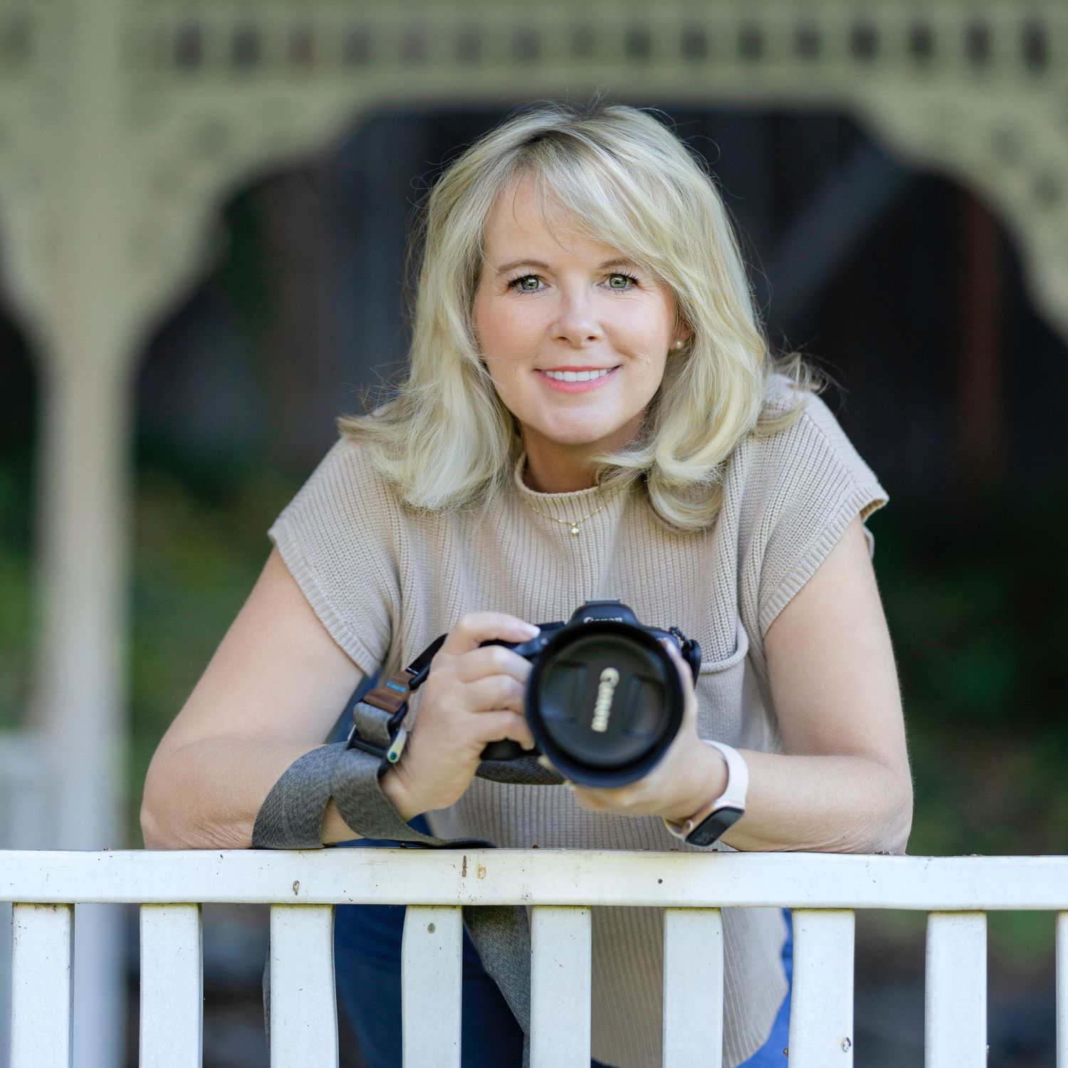 A photographer with blonde hair leans on a white railing while holding a professional DSLR camera.
