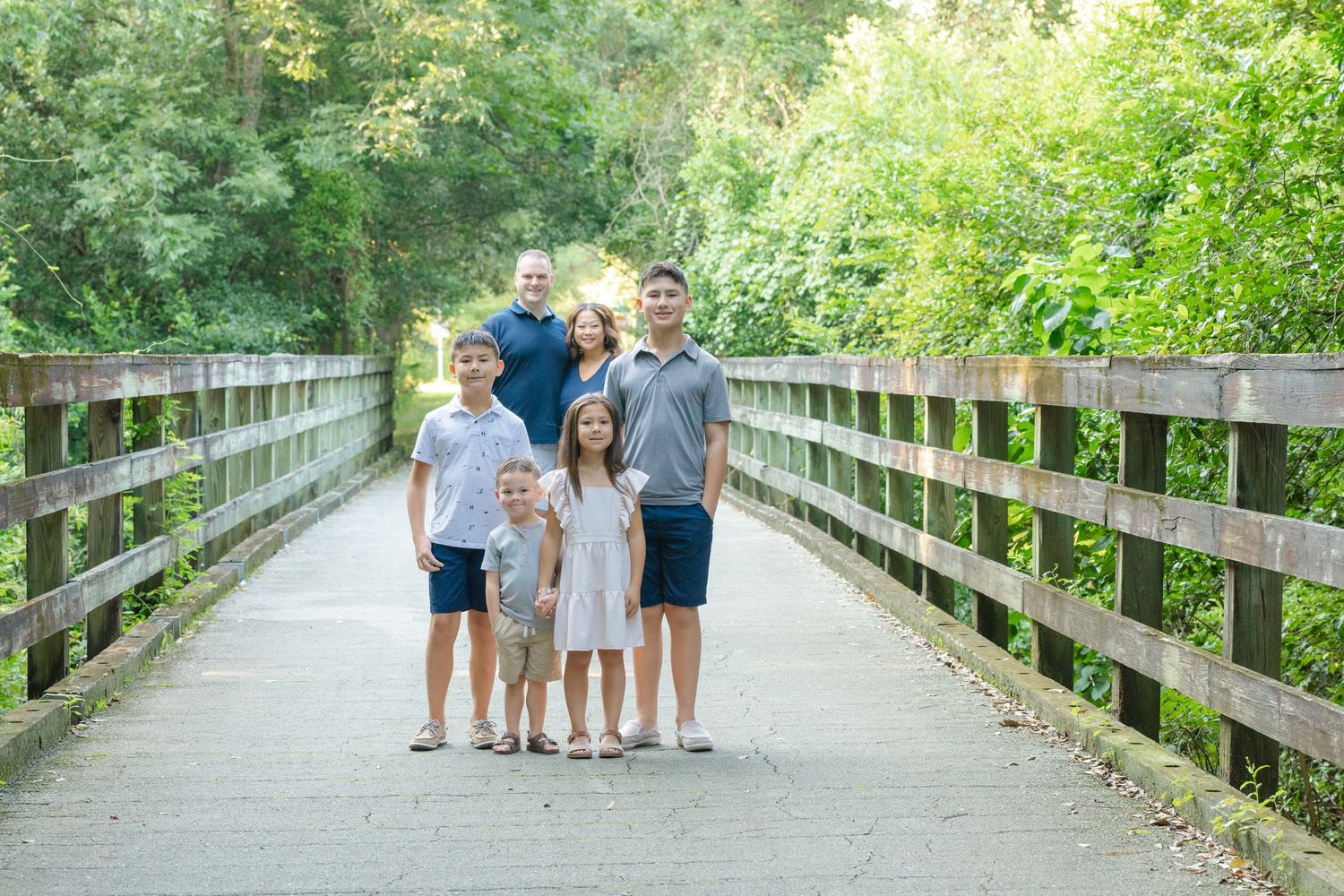 Family of six posing on a wooden bridge surrounded by lush green trees on a sunny day.