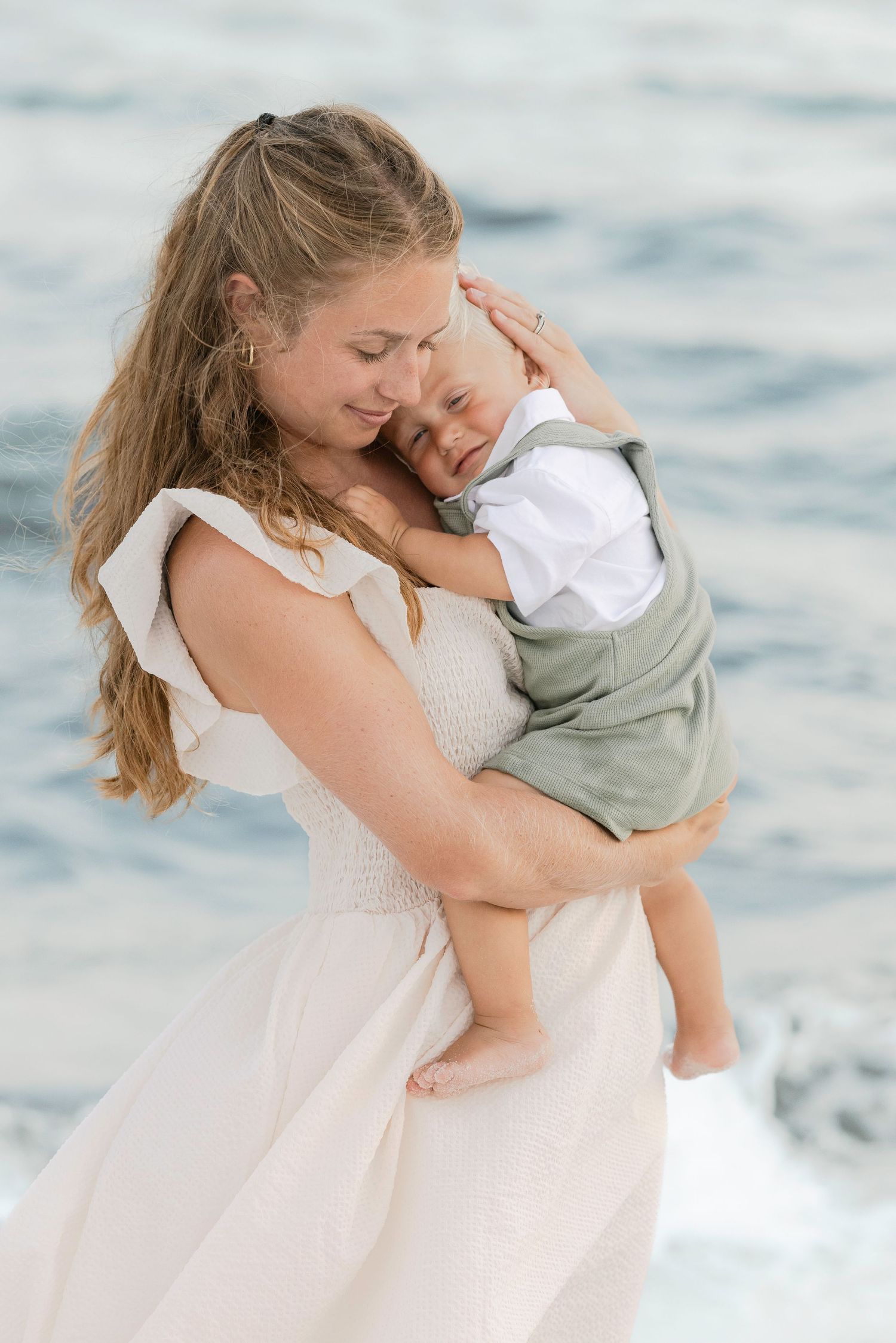 Loving mother in white dress cradling sleeping baby by the ocean waves.