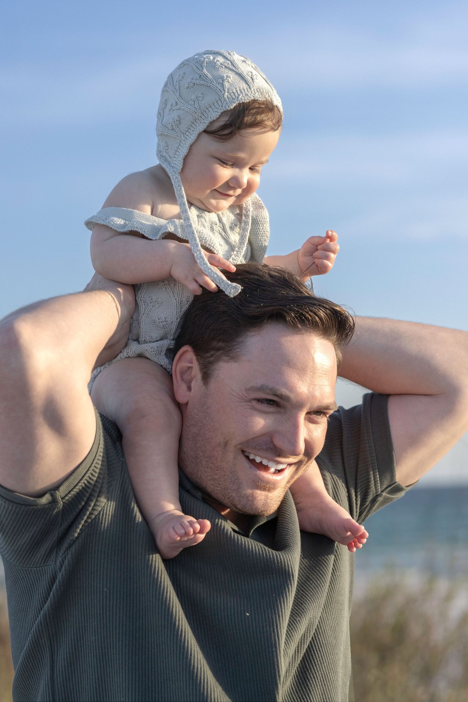 Smiling father carries happy baby in knit bonnet on his shoulders at a sunny beach.