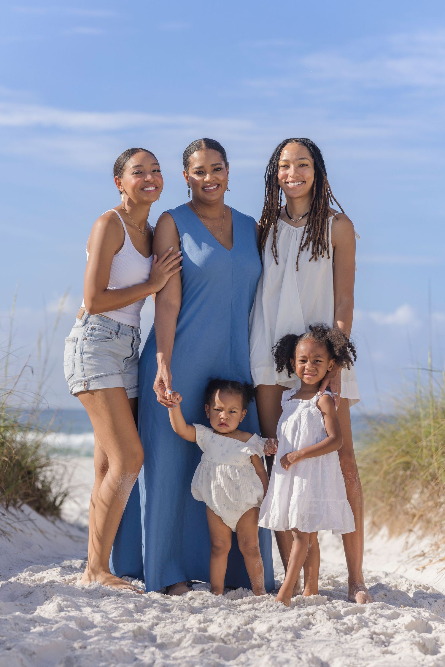 A joyful family of five poses on a sunny beach, with three women and two young girls dressed in white and blue.