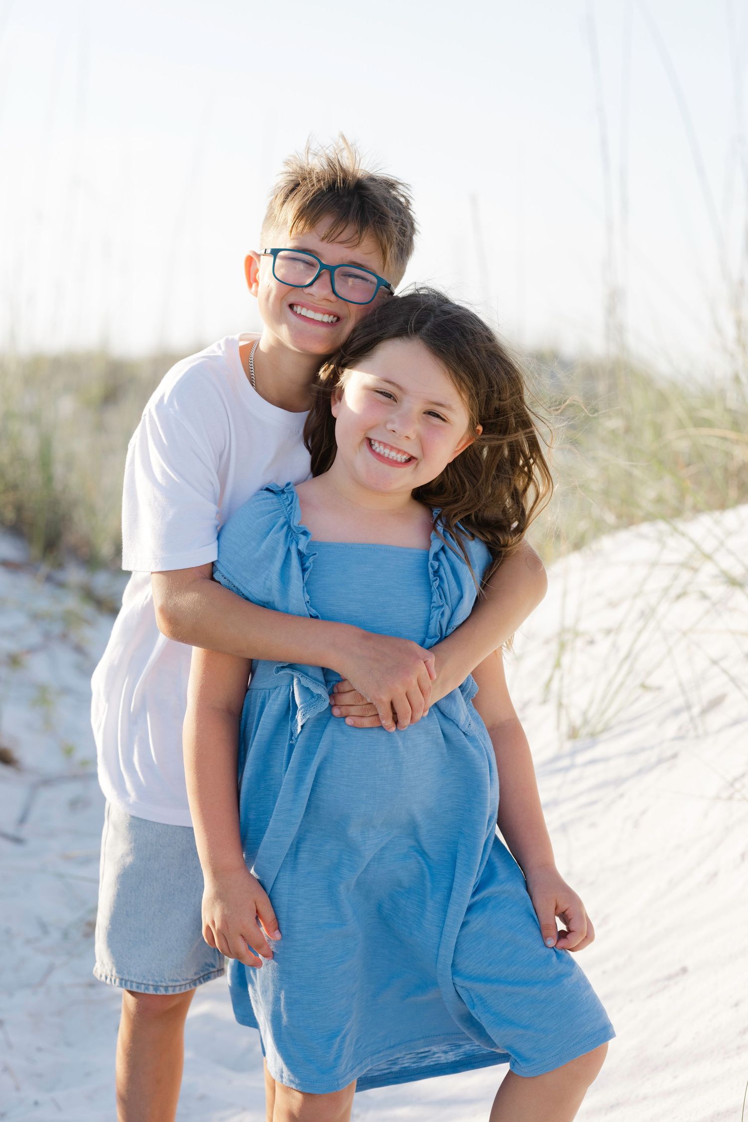 Two smiling children hugging on a sandy beach, boy in white shirt and glasses, girl in blue dress.
