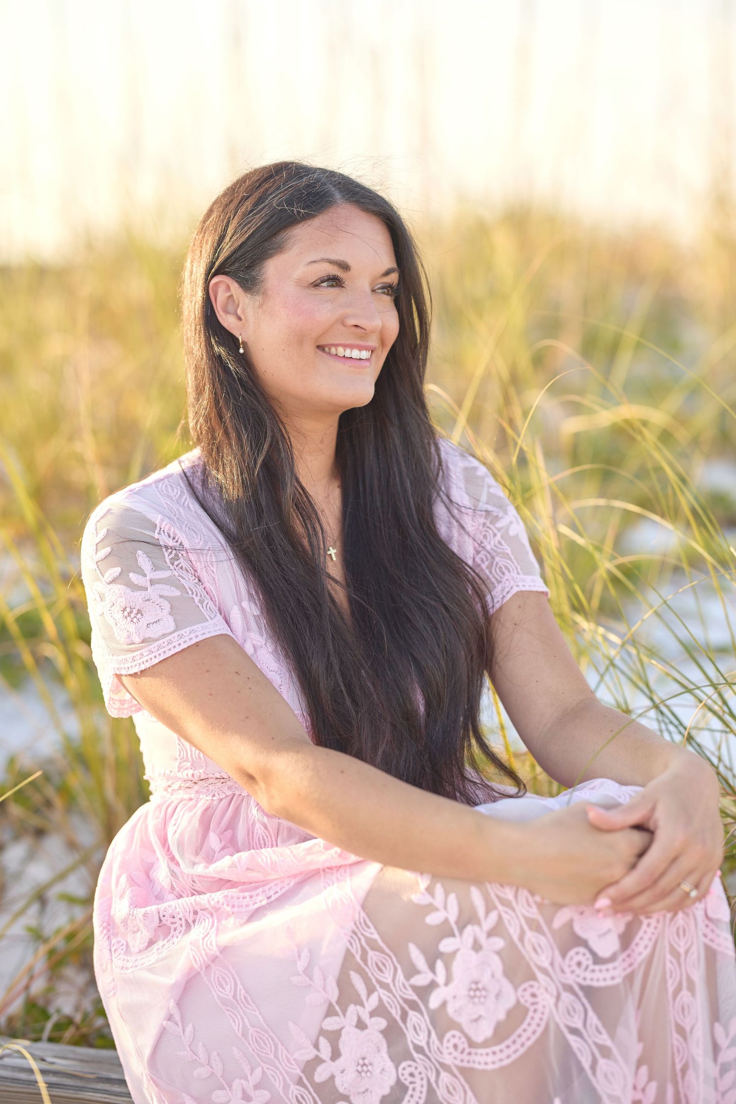 Smiling brunette woman in pink floral lace dress sitting on beach with sea grass and sand dunes behind her.