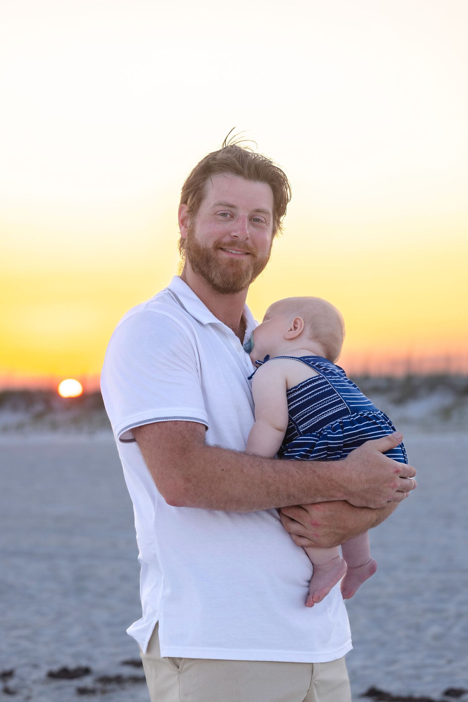 Bearded father in white polo shirt tenderly holding baby at beach during vibrant golden sunset.