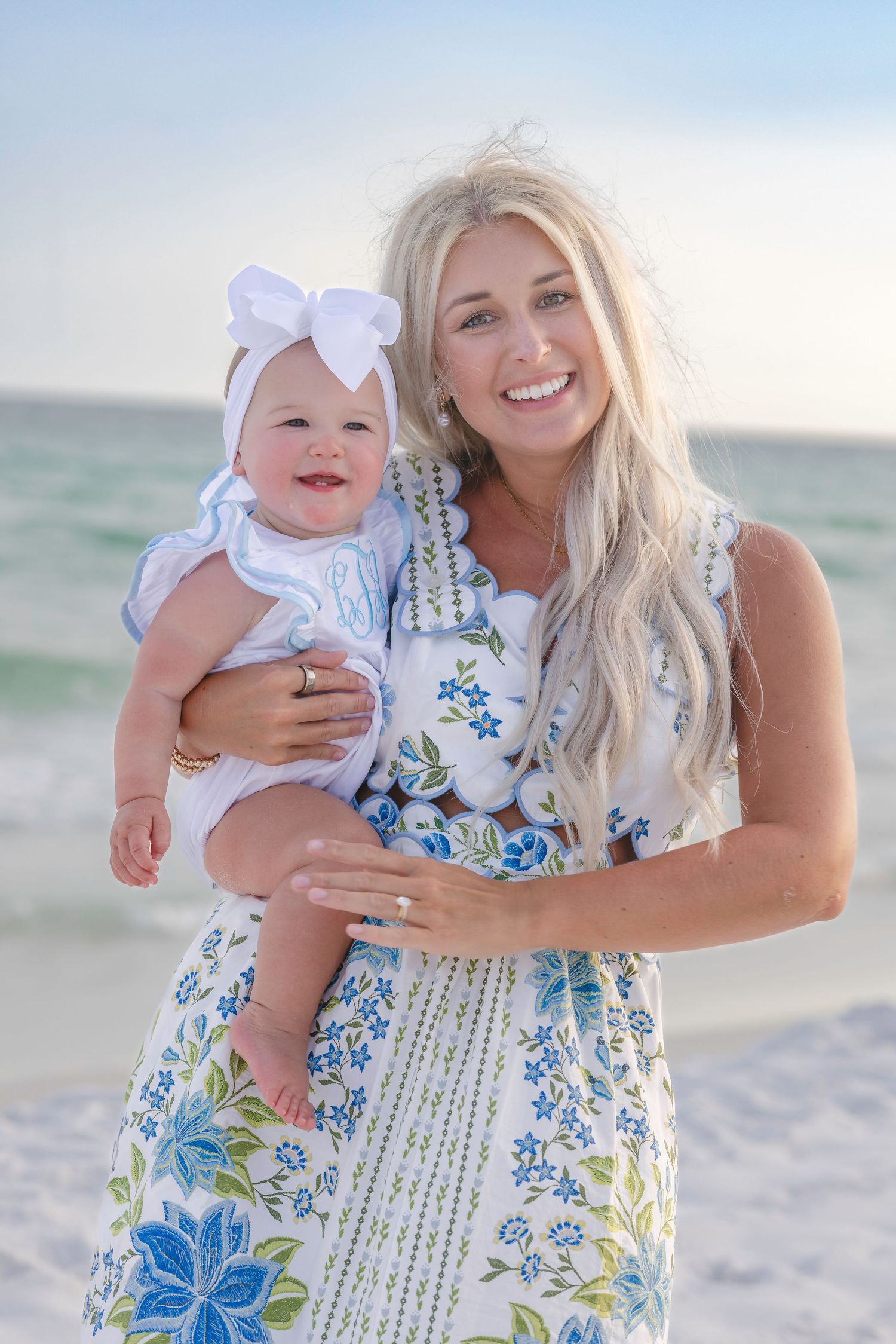 Smiling blonde woman in floral dress holds a happy baby girl with a white bow on a sunny beach.