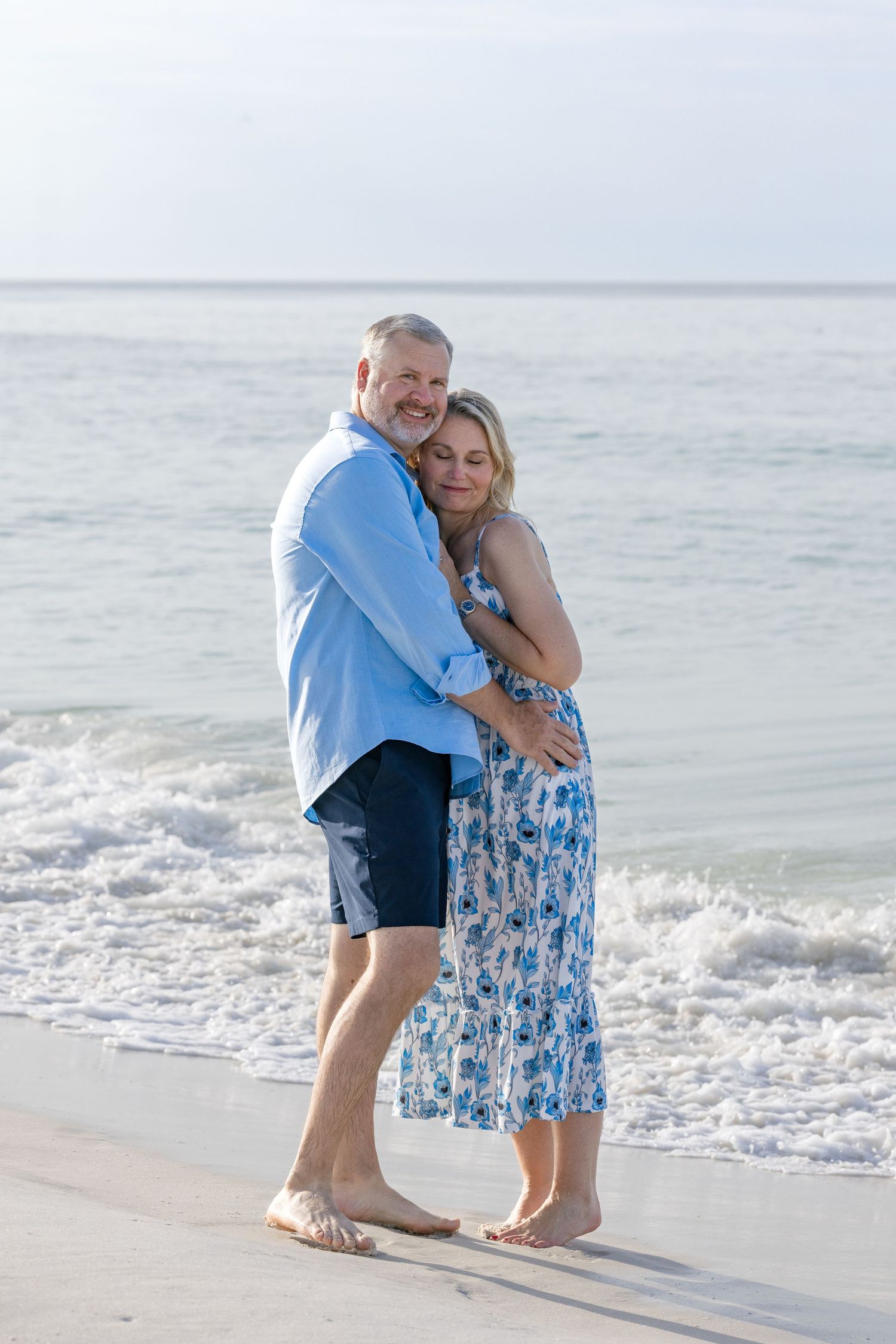 A couple embraces barefoot on a sandy beach, the man in blue shirt and shorts, woman in floral dress, ocean waves behind them.