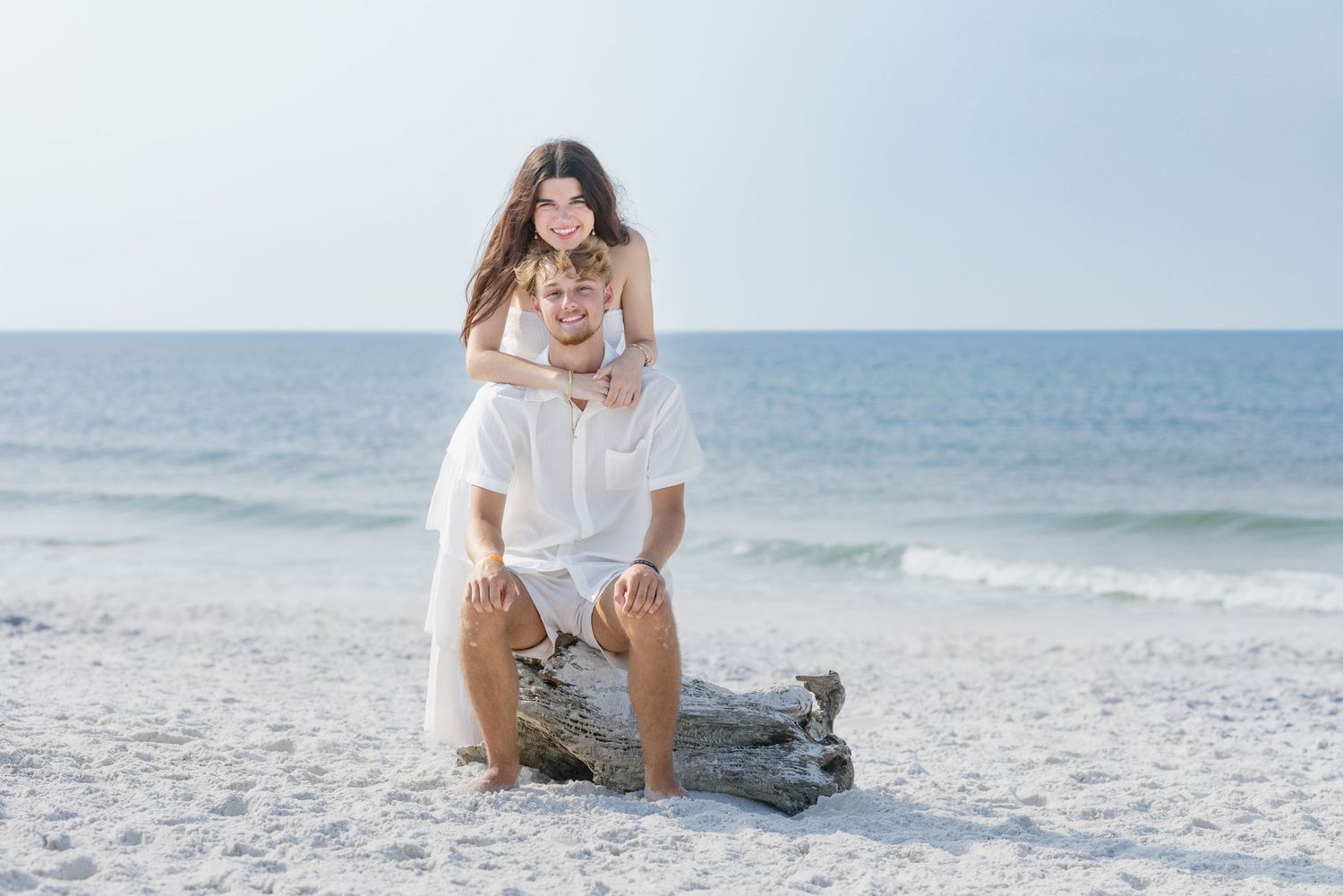 Couple posing on driftwood at a white sand beach, woman standing behind man, both wearing white outfits.