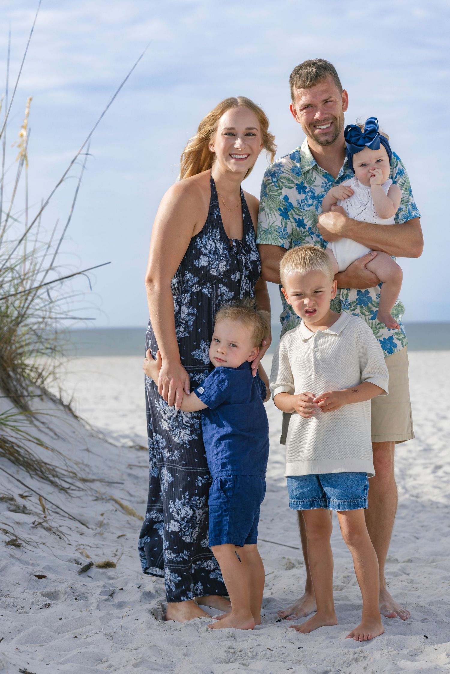 Happy family of five posing on a sandy beach, parents with three young children in coordinating blue outfits.