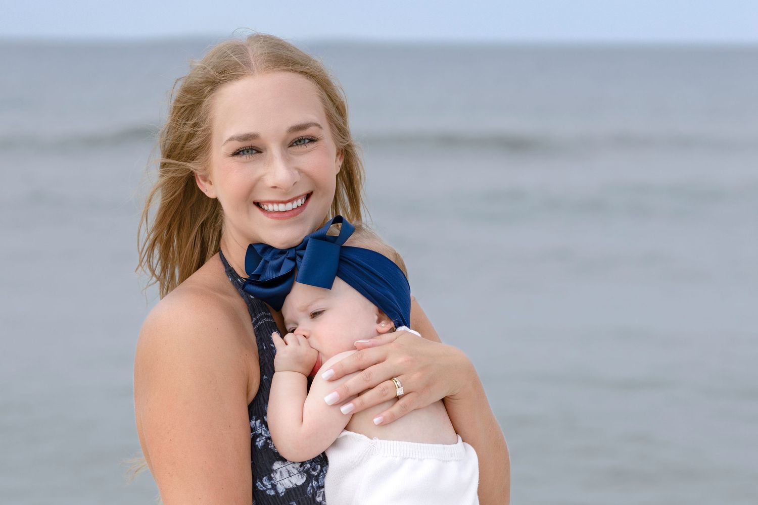 Smiling young mother holding sleeping newborn baby with blue bow on sunny beach.
