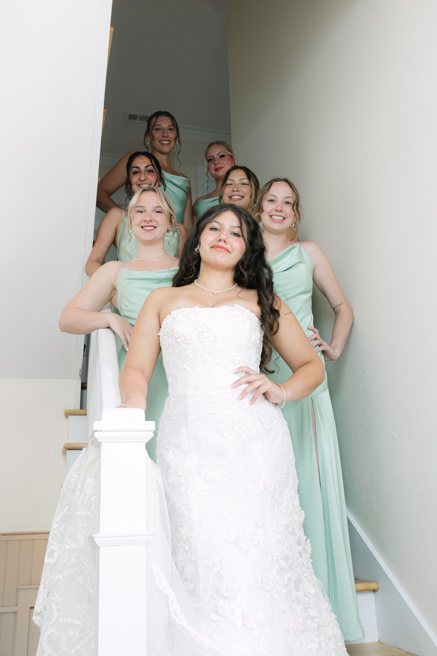 Bride in white lace gown poses with bridesmaids in mint green dresses on an elegant staircase.