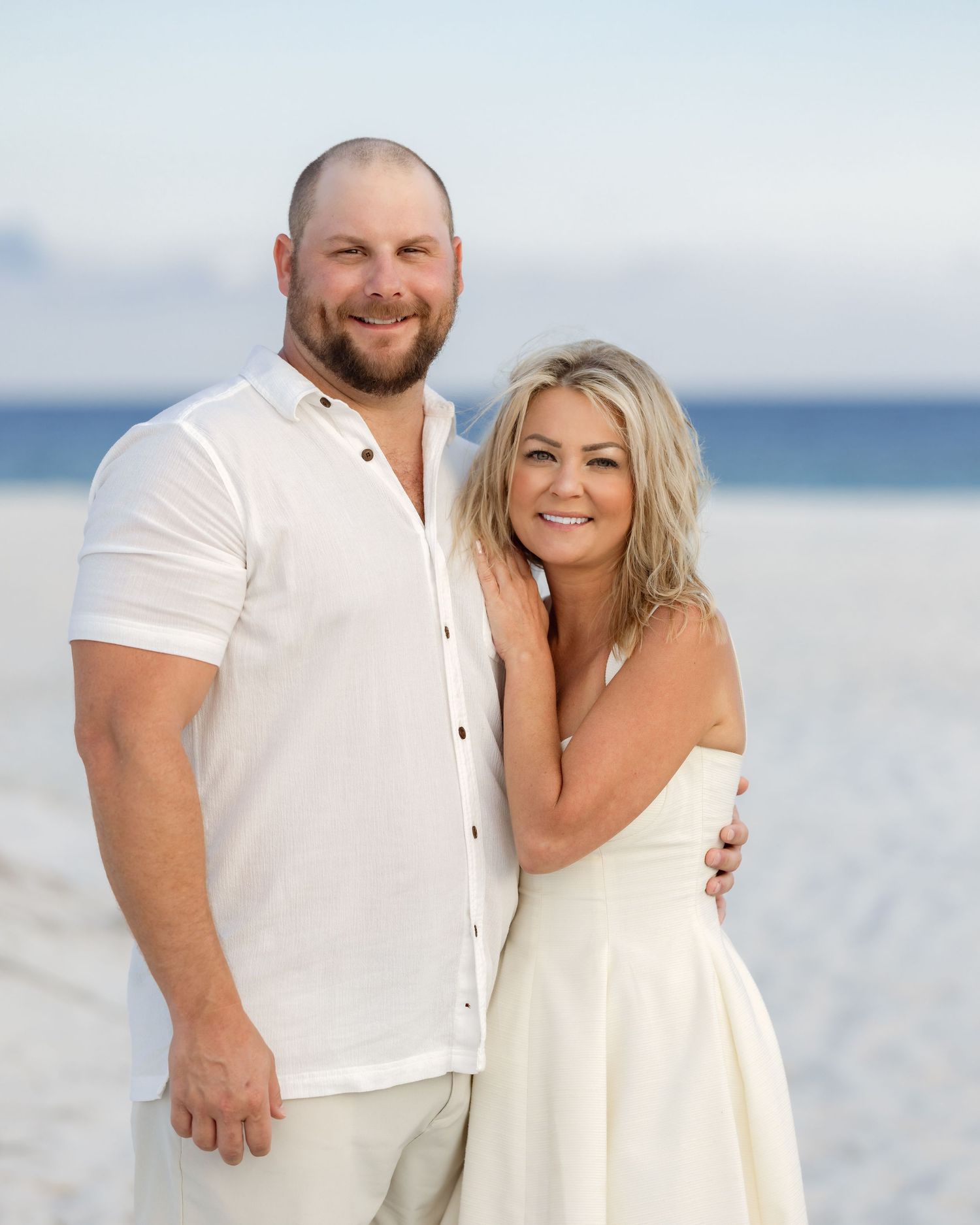 Smiling couple in white outfits embracing on sunny beach with ocean backdrop.