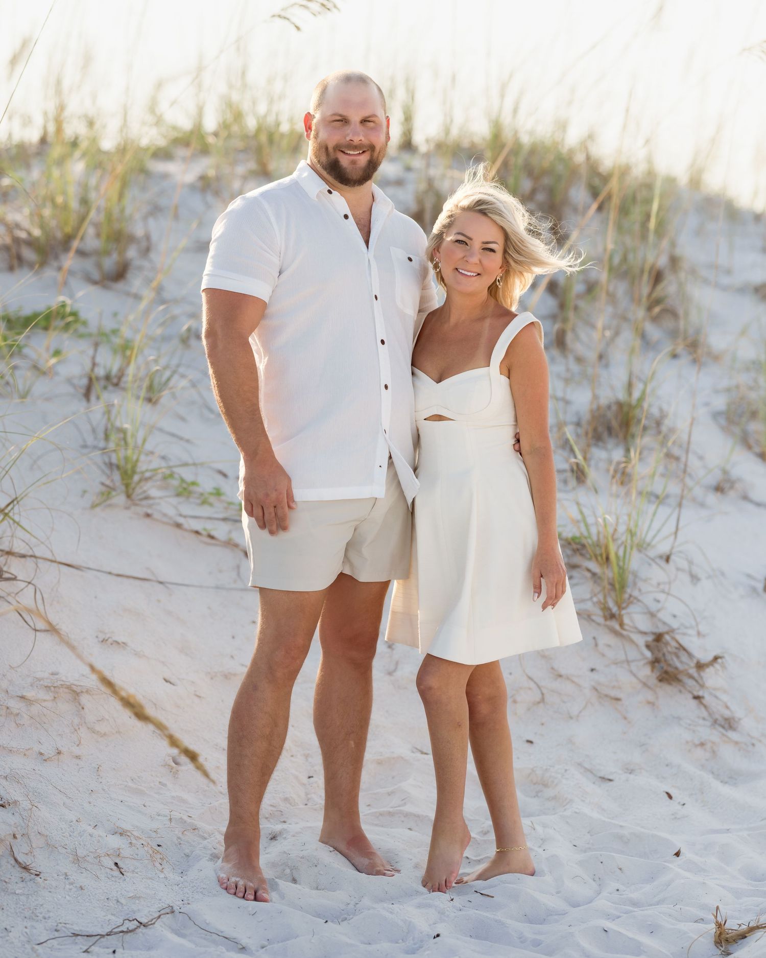 Couple in matching white outfits posing barefoot on sandy beach dunes with sea grass in background.
