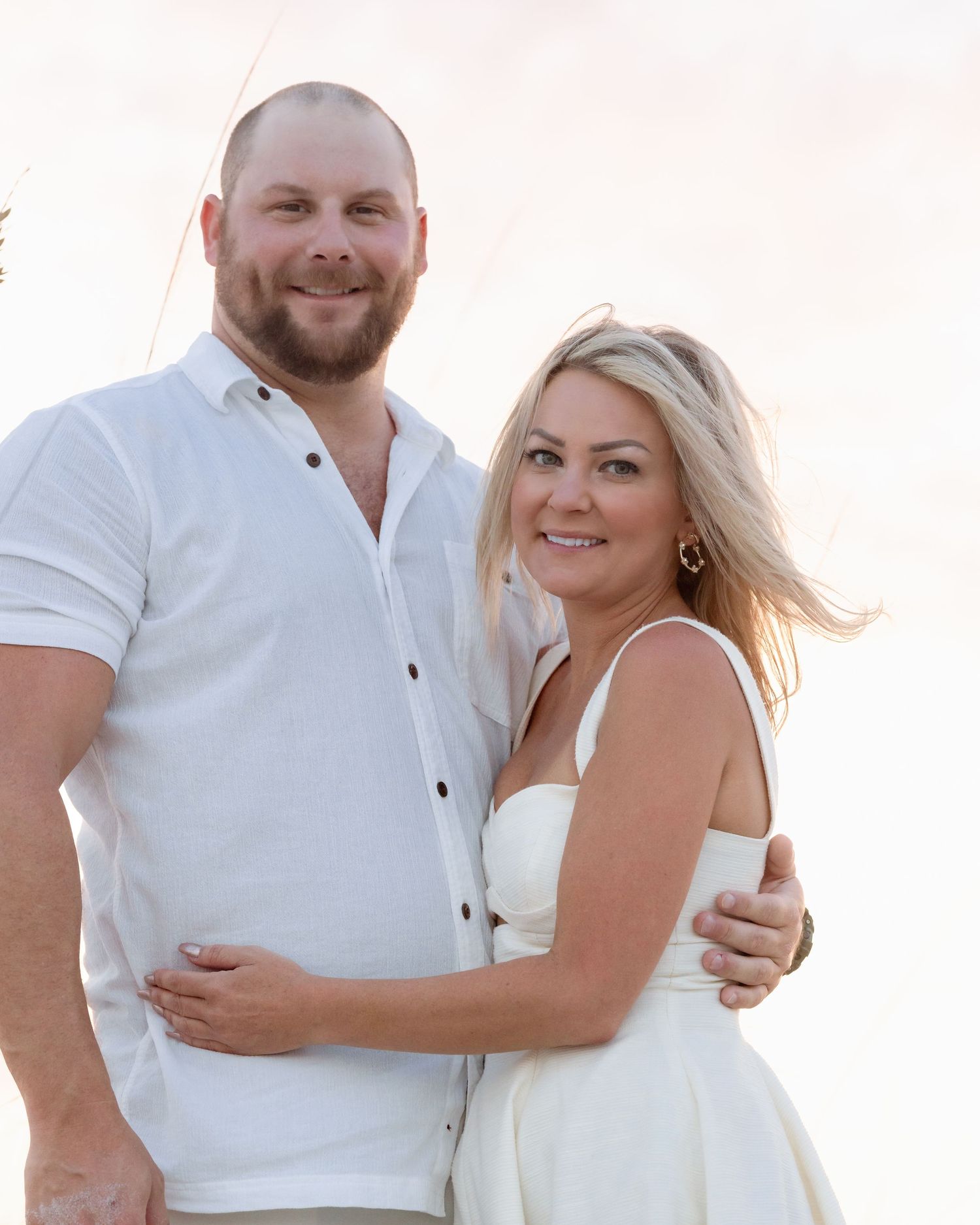 Smiling couple in matching white outfits embracing at golden hour on a beach.