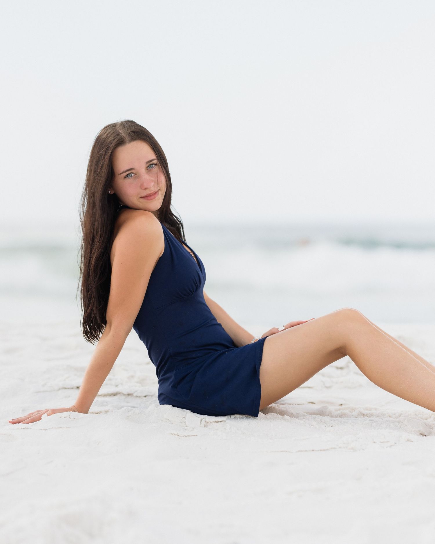 Young woman in navy blue romper sitting on white sand beach smiling at camera.