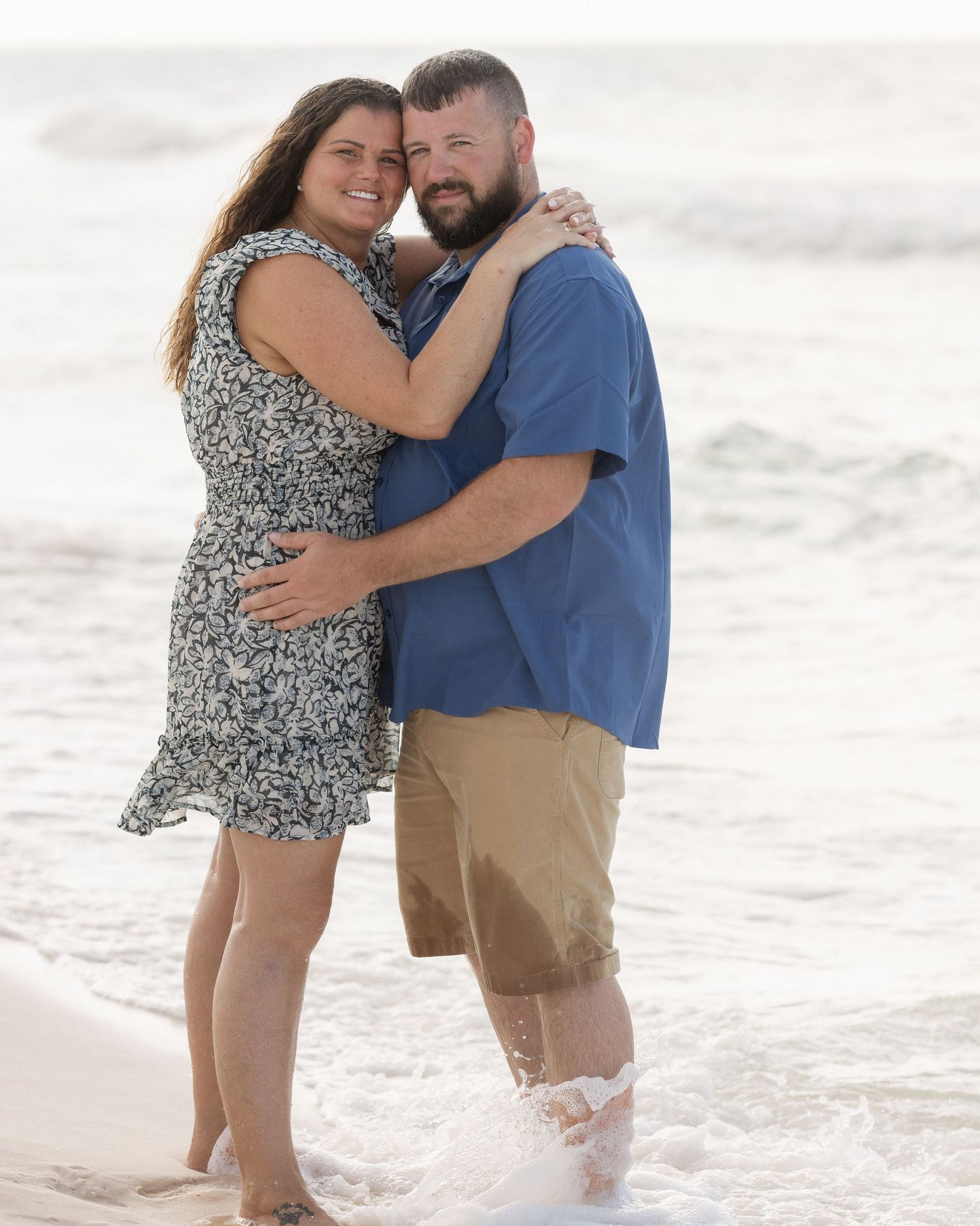 Couple embracing and smiling on a sandy beach with ocean waves, woman in floral dress and man in blue shirt.