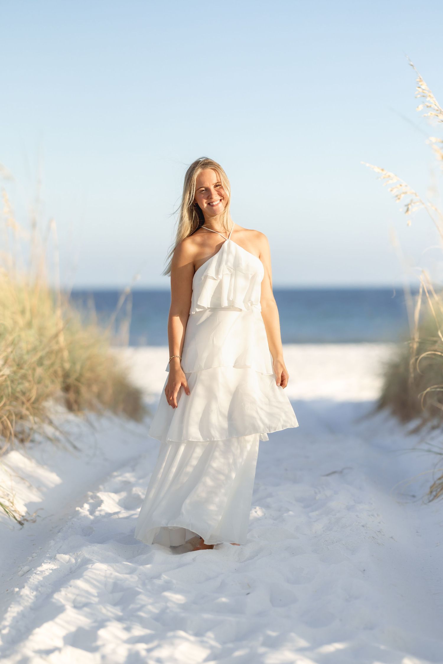 Blonde woman in white tiered ruffle maxi dress posing on sandy path between dunes with ocean in background.
