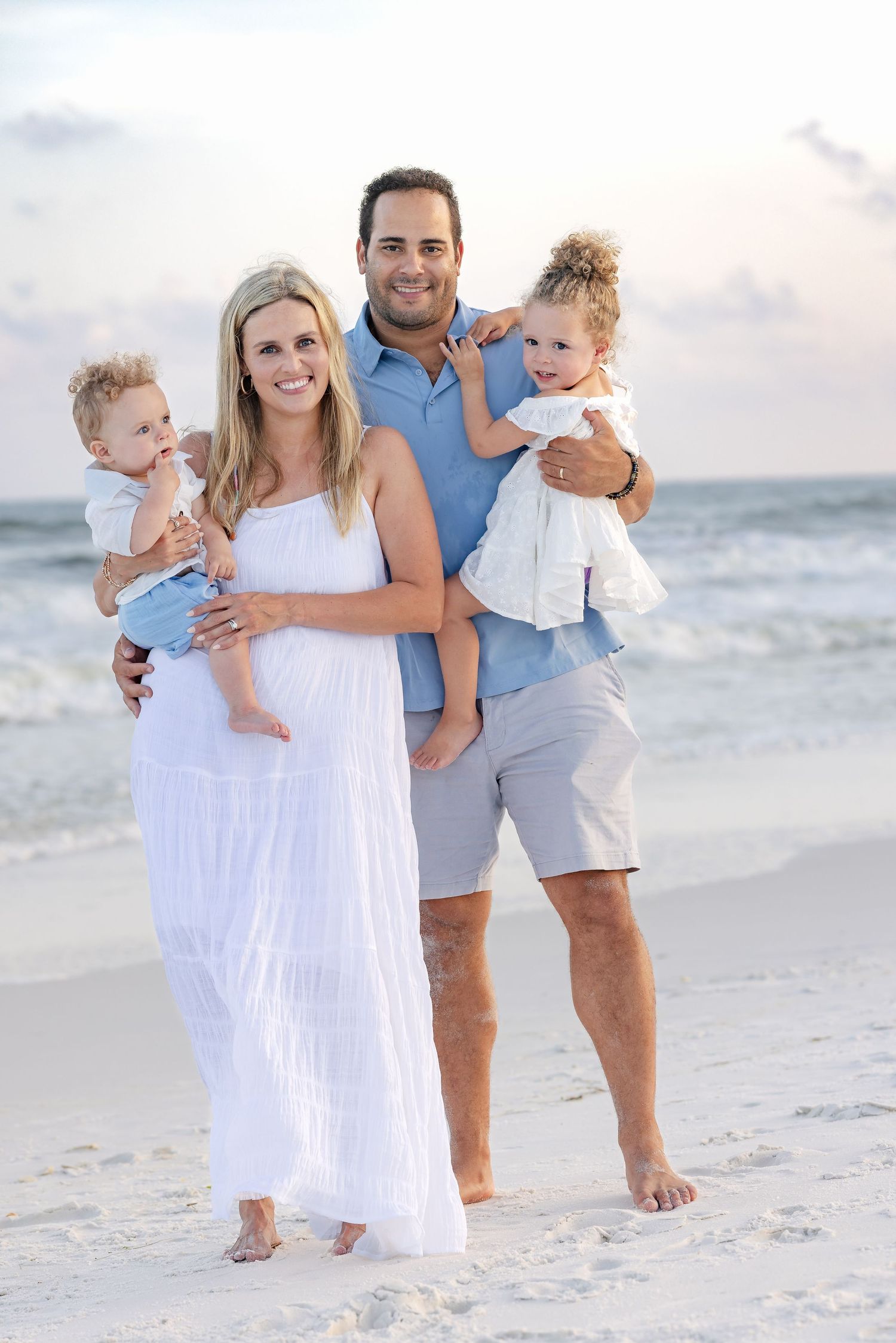 Happy family of four posing on a sandy beach at sunset, parents holding two toddlers, wearing white and blue outfits.