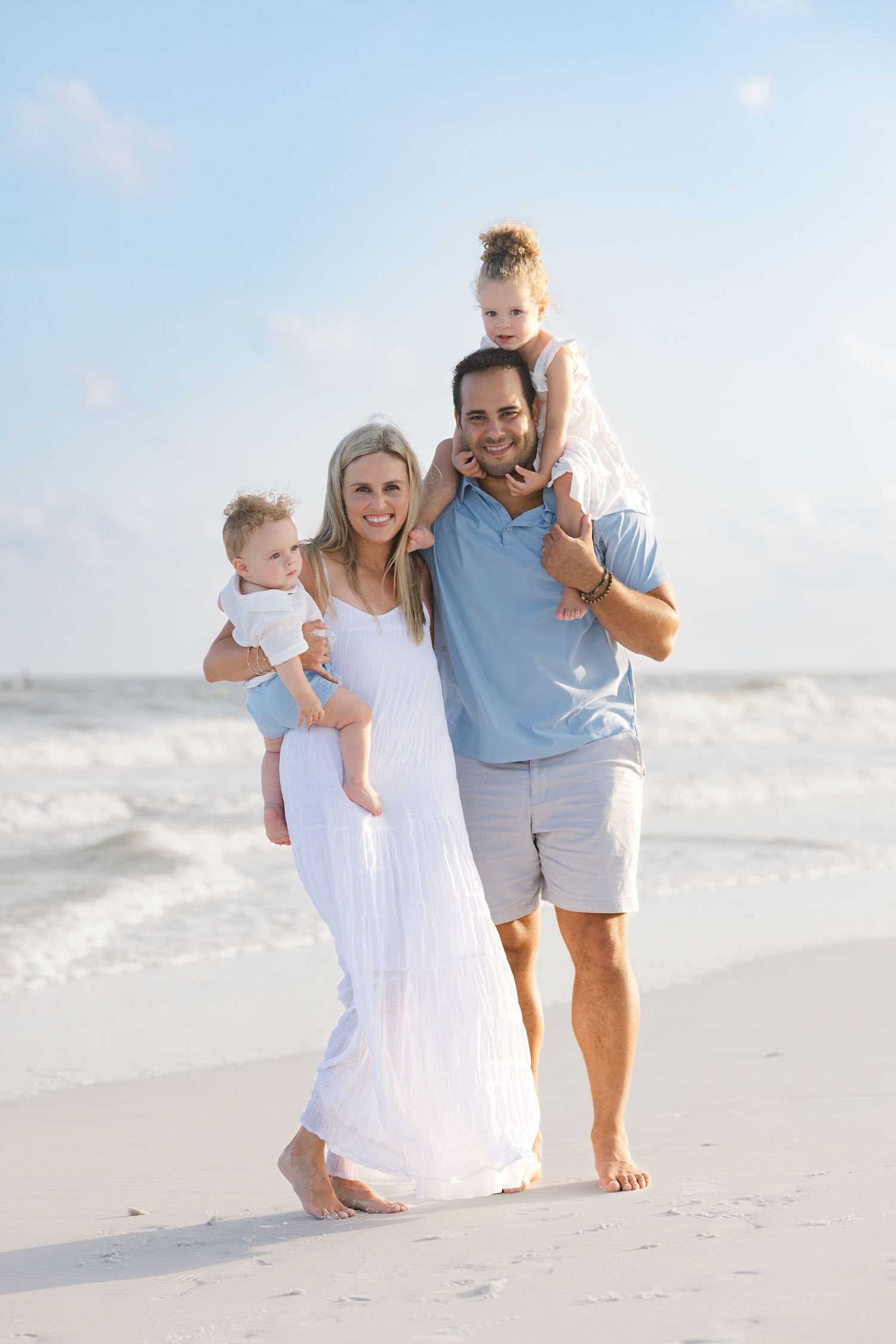 Happy family of four posing on a sunny beach, parents holding two young children with ocean waves in the background.