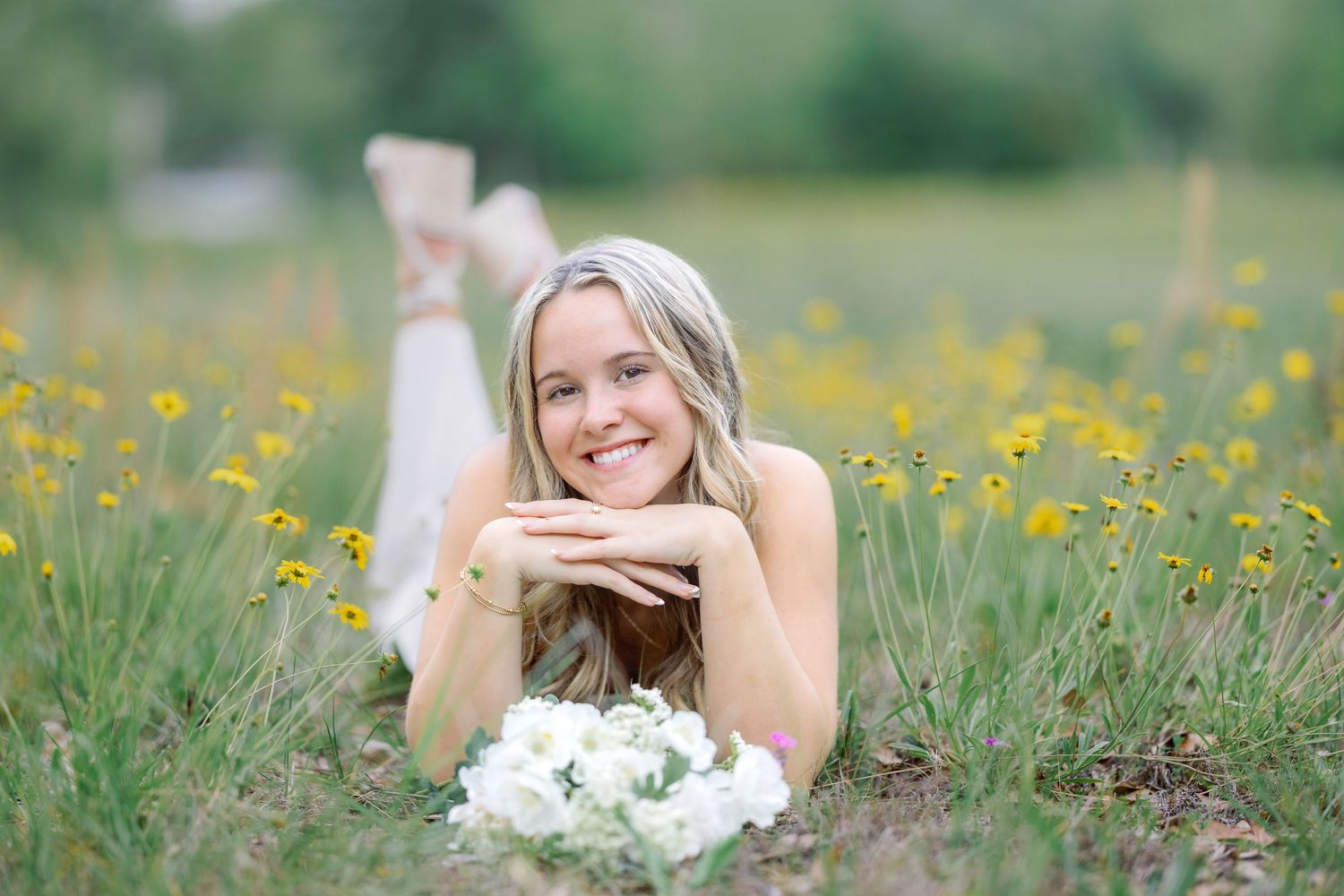 Smiling blonde woman lying in a wildflower field with white flowers, surrounded by yellow blooms in a lush green meadow.
