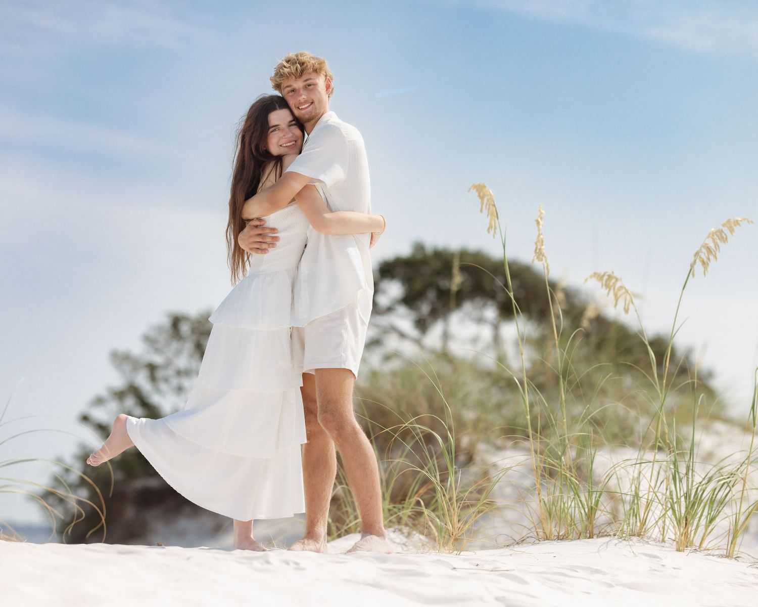 Couple embracing in white outfits on white sand dunes with sea oats and blue sky.