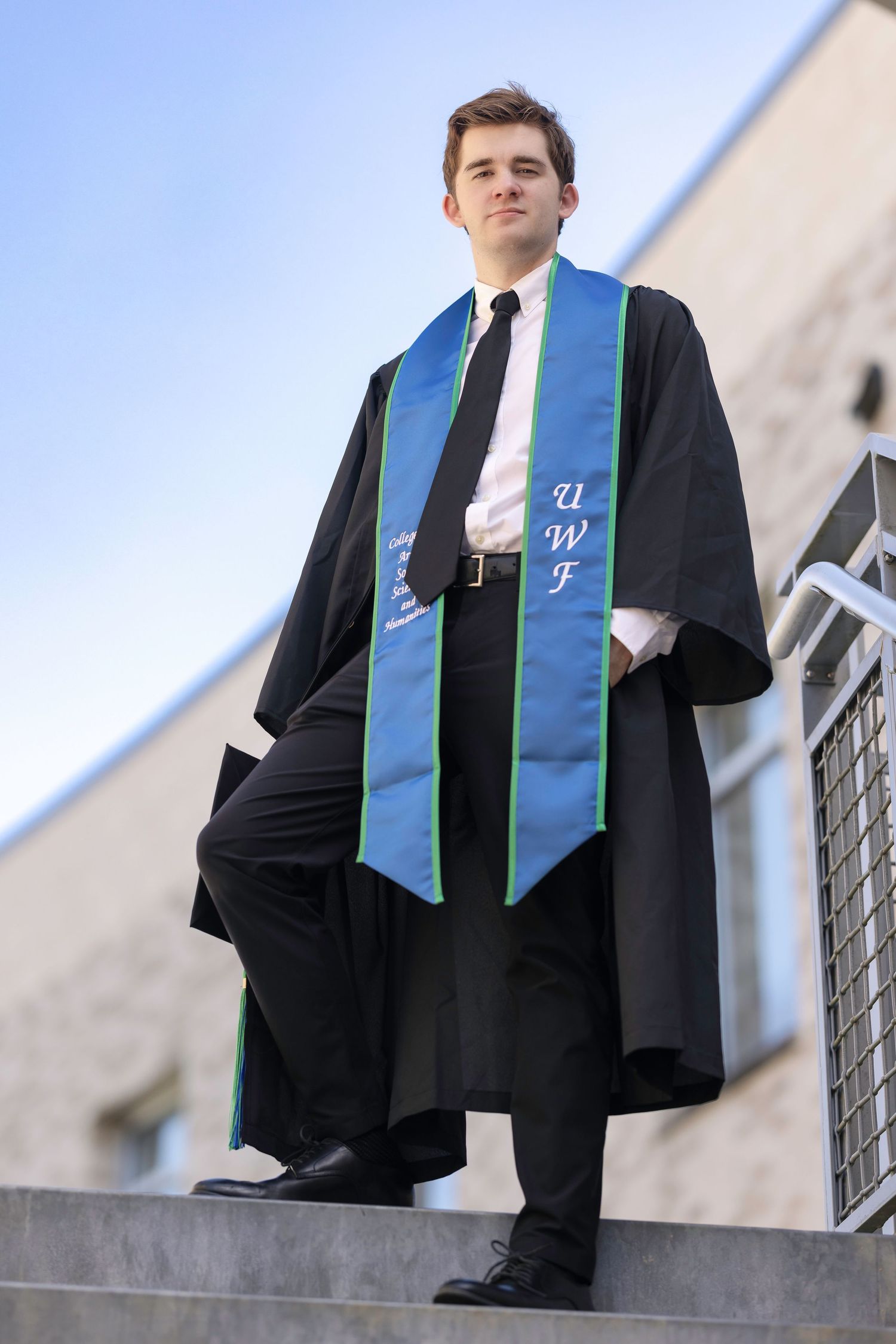 Young man in black graduation gown with UWF blue stole standing proudly on steps outdoors.