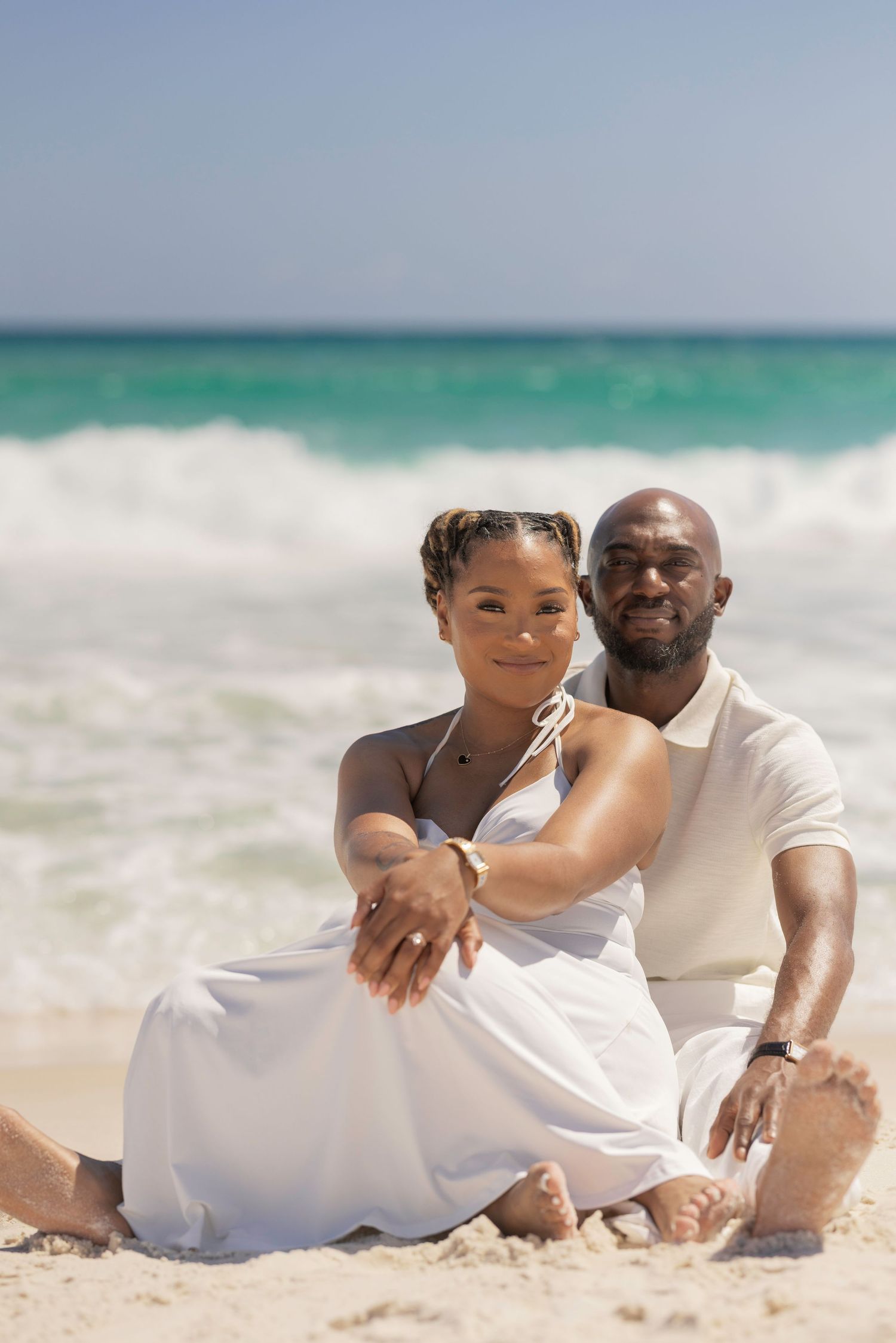 A smiling couple in white outfits sits together on a sandy beach with turquoise ocean waves behind them.