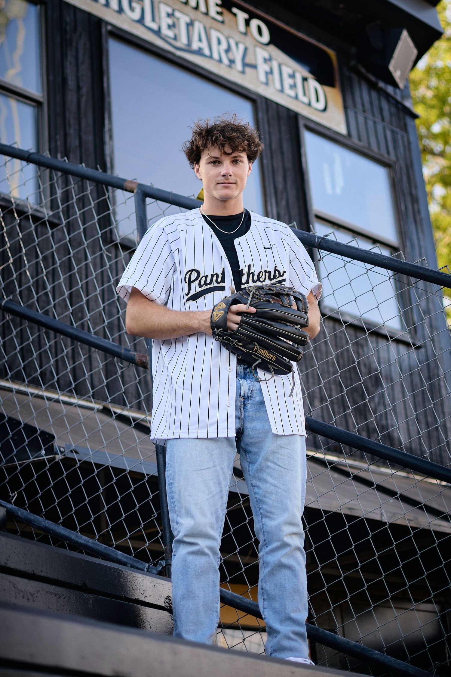 Teen baseball player in Panthers pinstripe jersey holds glove at Singletary Field stadium bleachers.