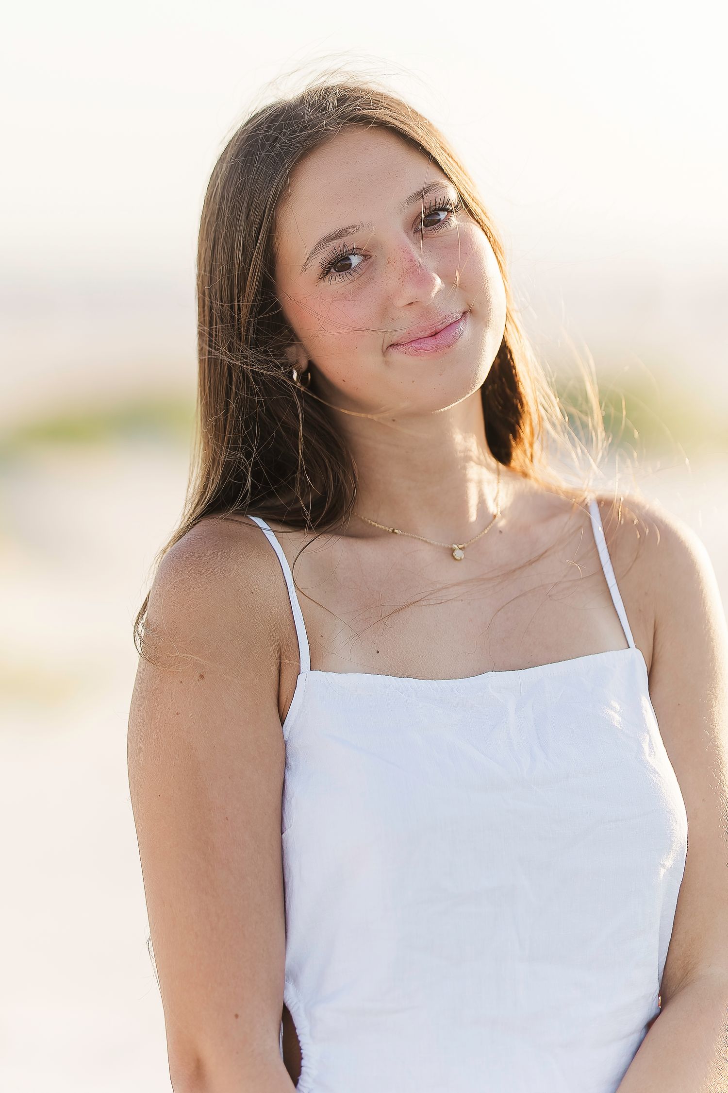 A person in a white tank top poses against a bright sunny background at the beach.